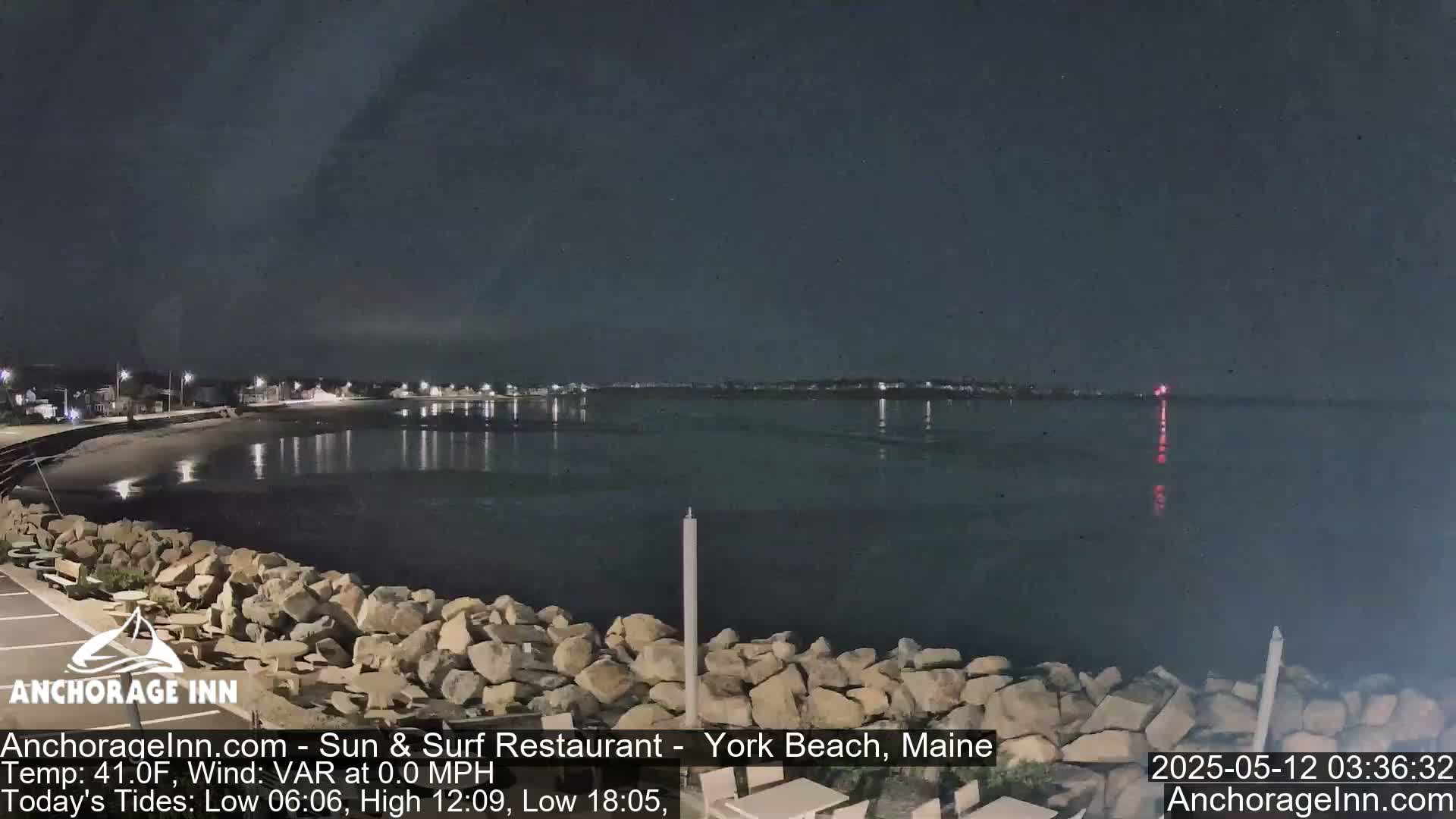 A nighttime view of a rocky shoreline and calm water, with lights visible in the distance across the water.