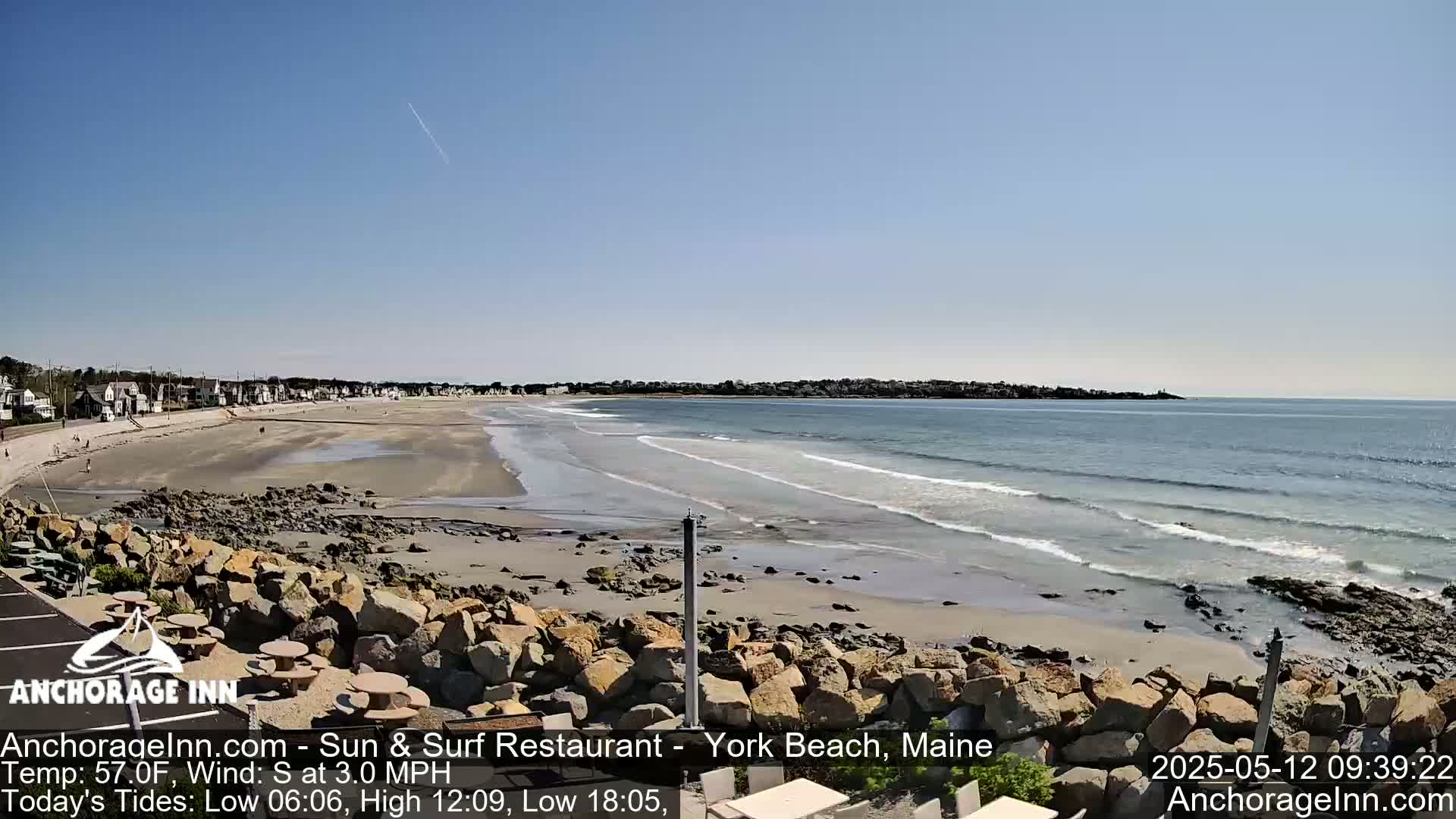 A sandy beach with gentle waves under a clear blue sky stretches out to a calm ocean, backed by a row of houses.