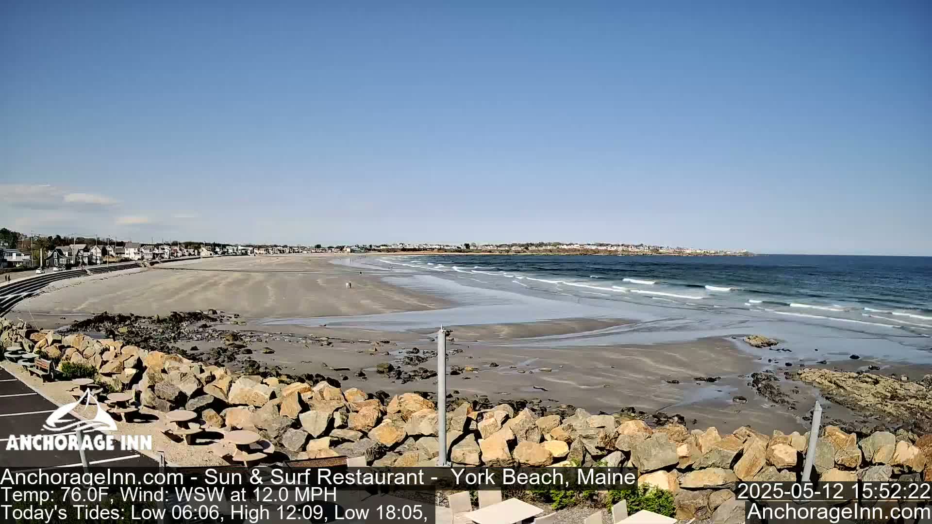 A sandy beach with small waves under a clear blue sky stretches out to a town on the horizon, with a rocky embankment in the foreground.