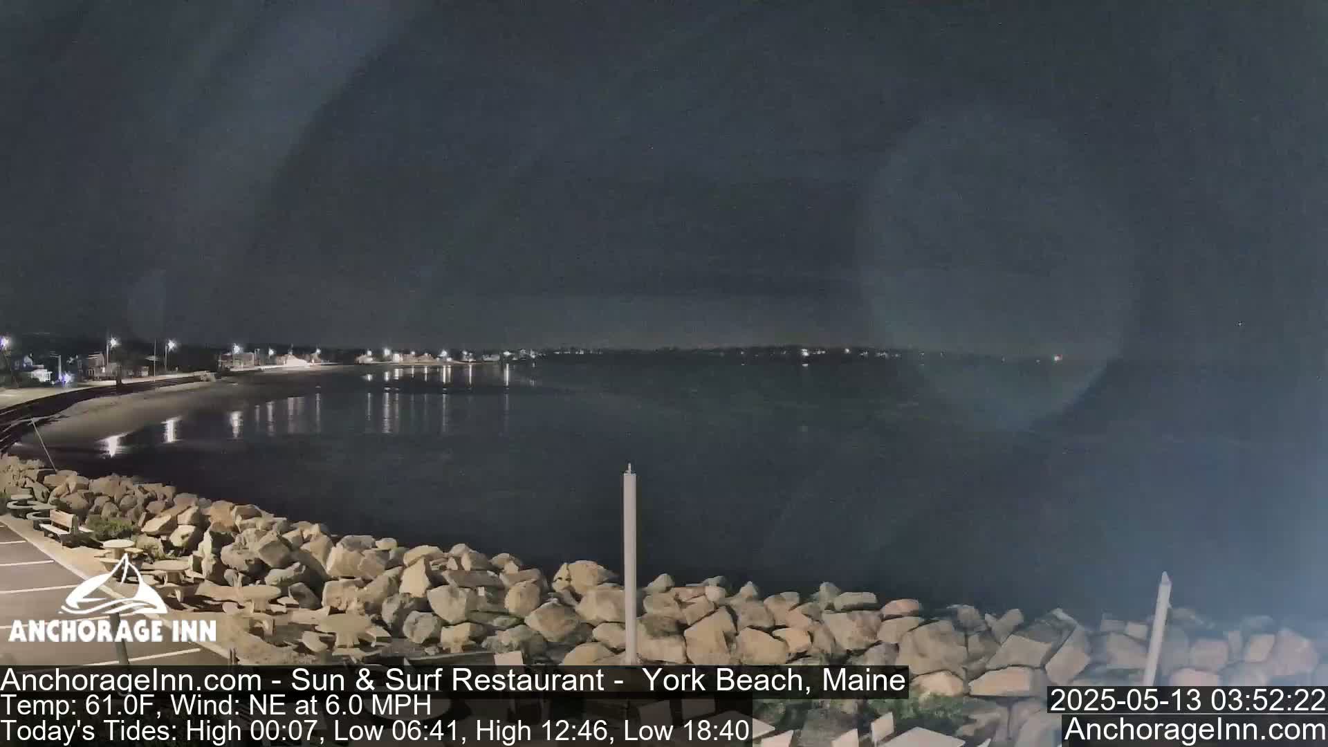 A nighttime view of a calm body of water next to a rocky shoreline, with a town faintly visible in the distance under a dark sky.