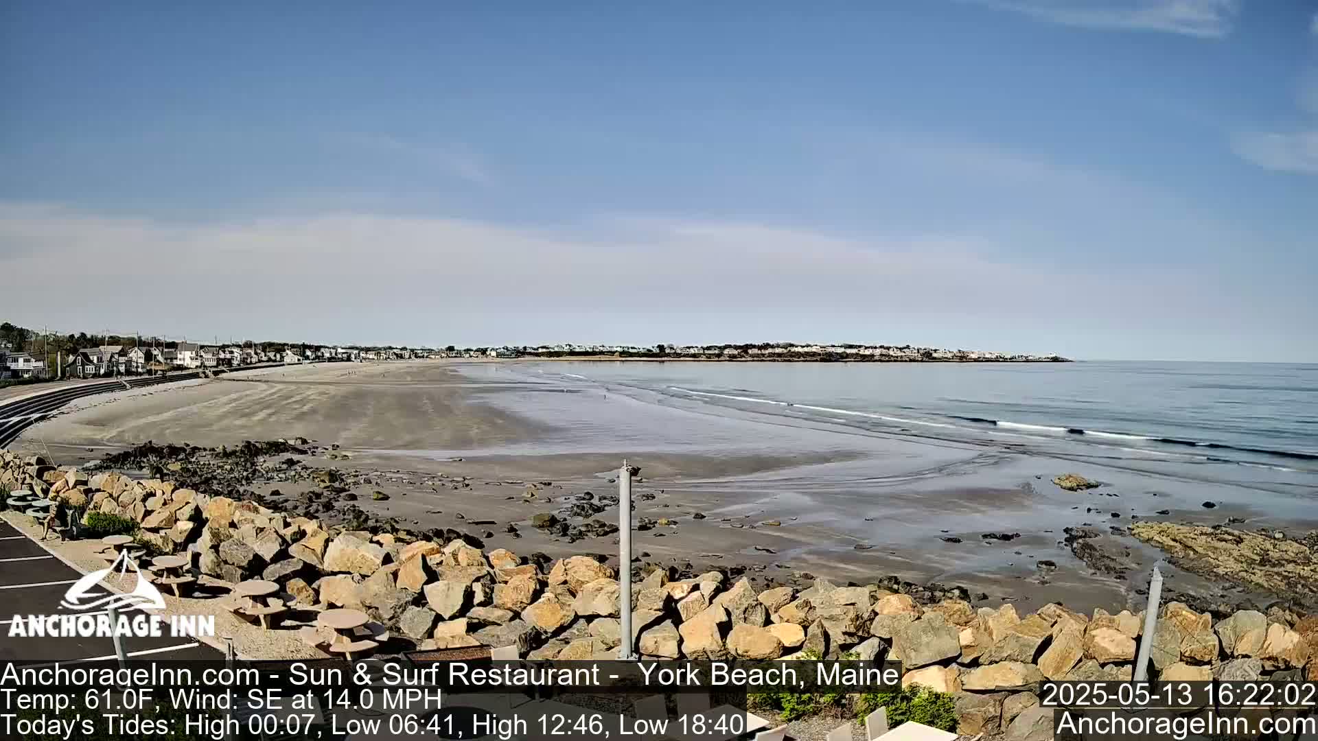 A sandy beach stretches along a calm ocean under a mostly clear sky, with a rocky barrier separating the beach from a viewing area.