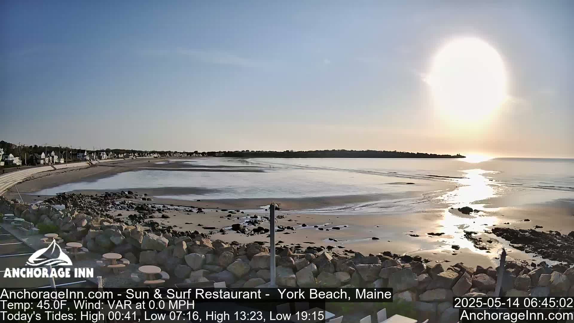 A sandy beach with calm water stretches out to a distant shoreline under a bright, hazy sun.