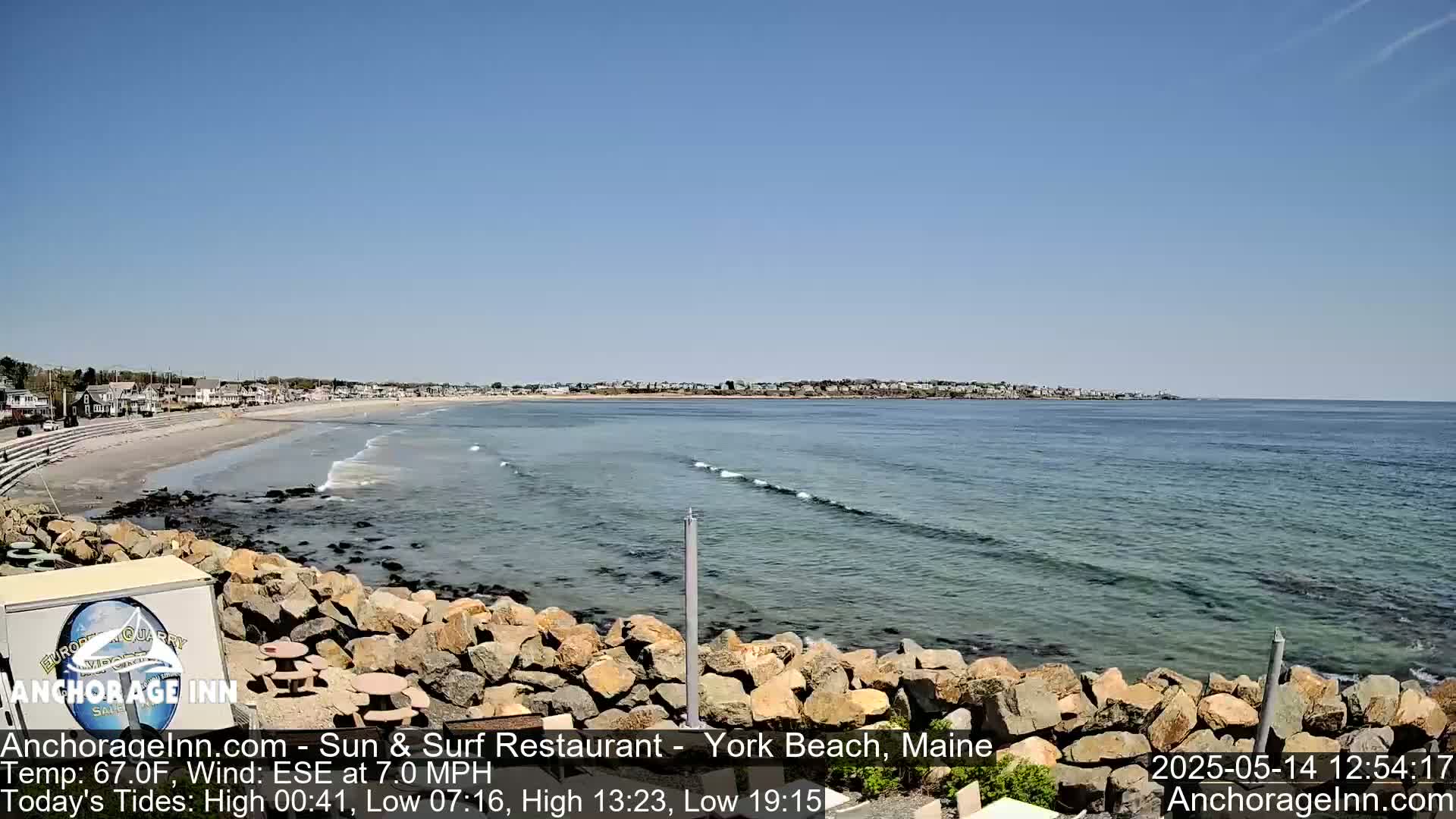 A sandy beach with calm waves and a clear blue sky is viewed from behind a rocky outcrop, showing a coastal town in the distance.