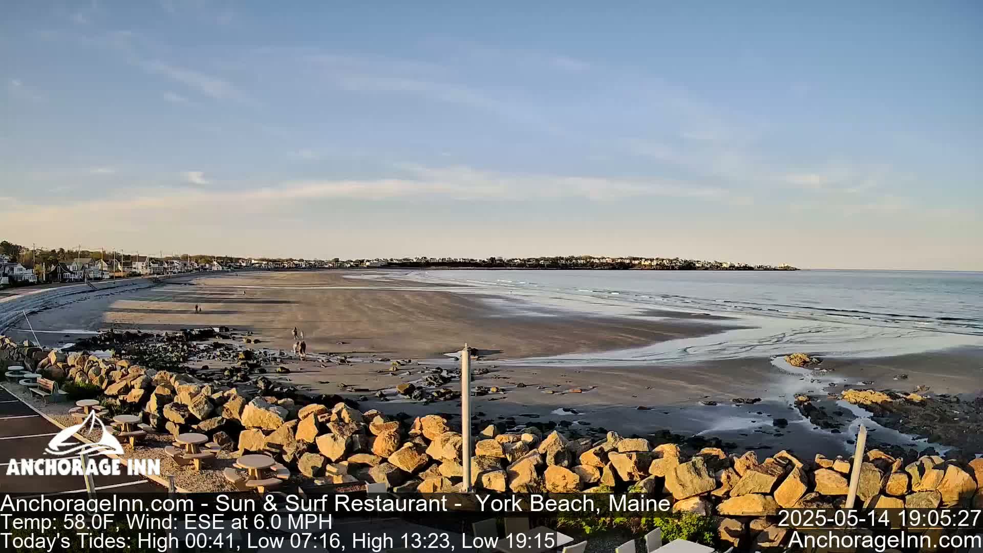 A wide sandy beach stretches to a calm ocean under a mostly clear sky, with a rocky barrier in the foreground and a small town visible in the distance.
