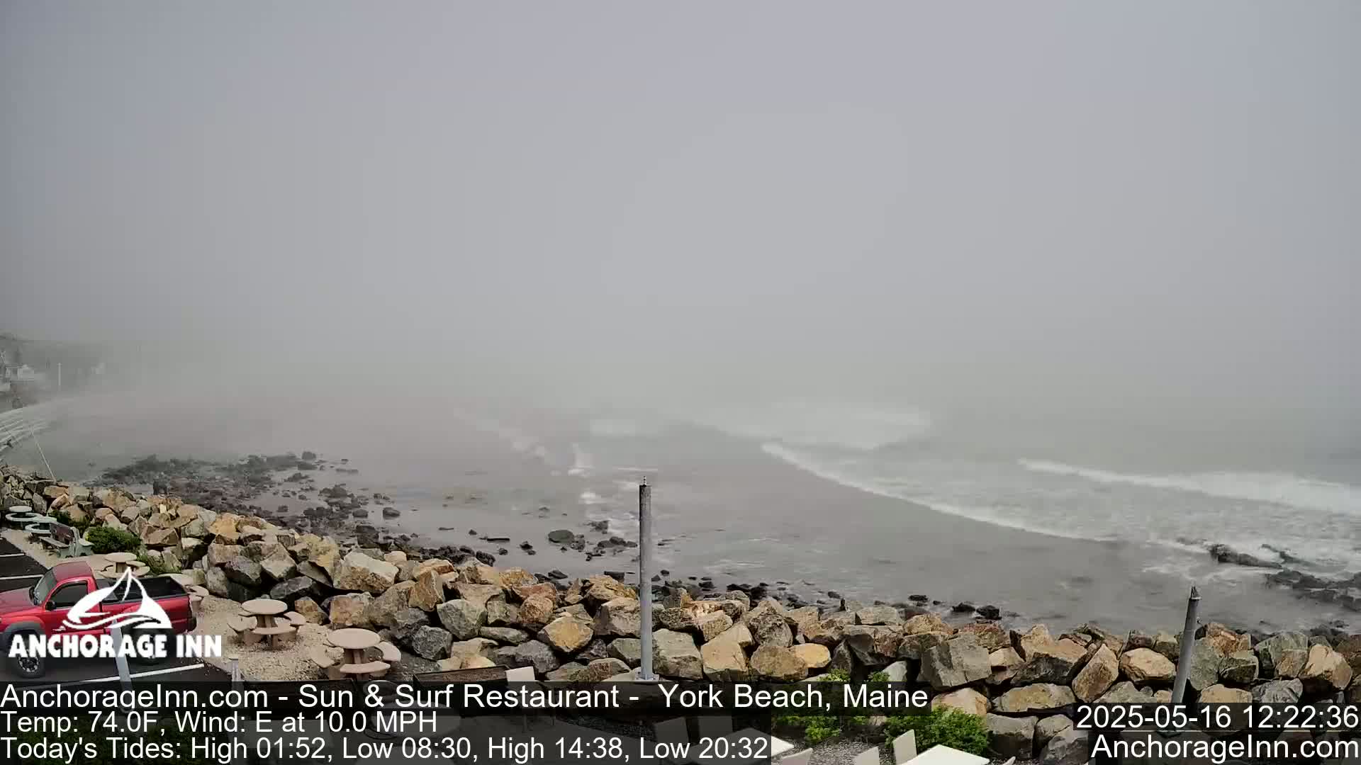 A foggy beach scene shows a rocky shoreline with gentle waves rolling in under a hazy sky.