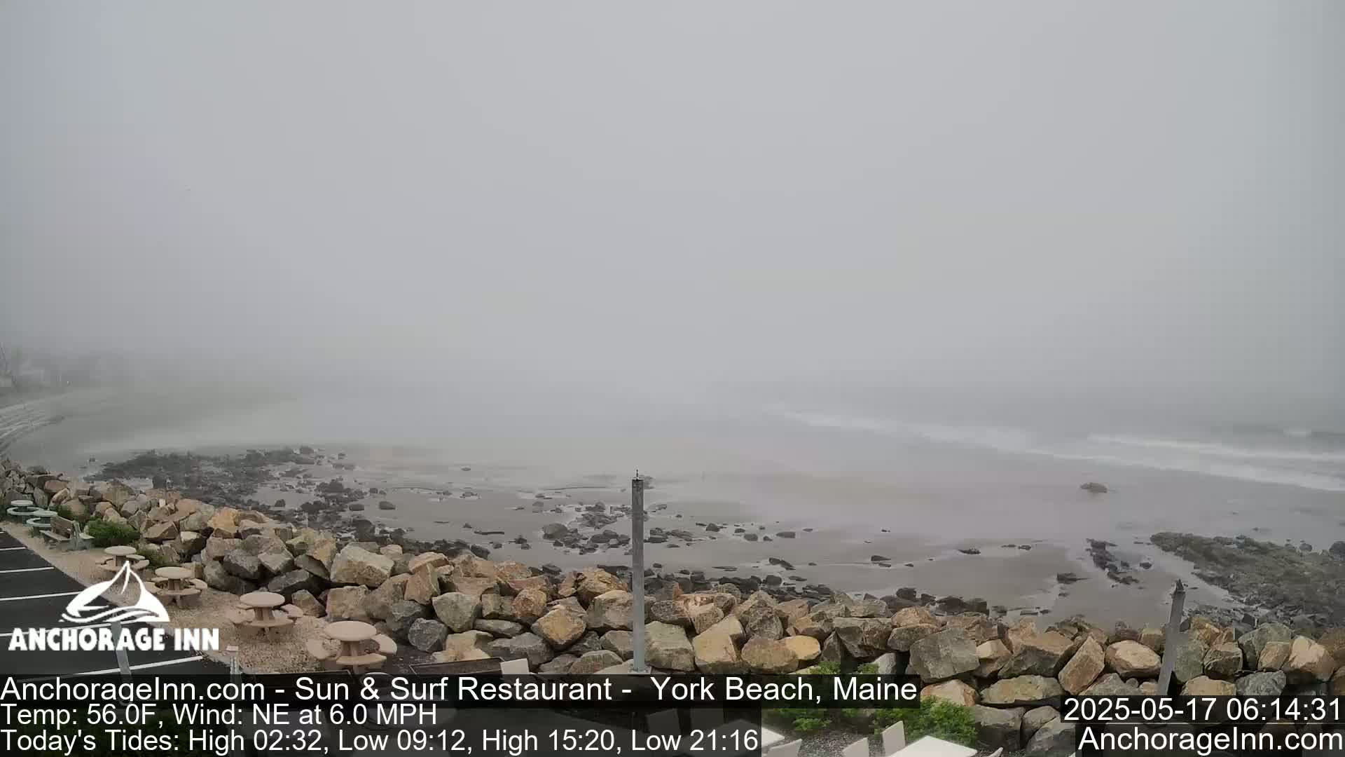 A foggy beach scene shows a rocky shoreline, gentle waves, and a mostly overcast sky.