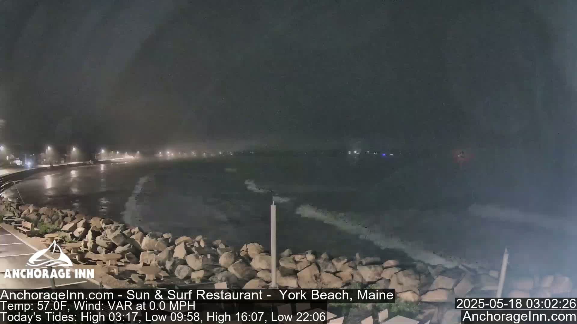 A nighttime, foggy scene shows a rocky shoreline with waves gently lapping against it, under a dark sky.