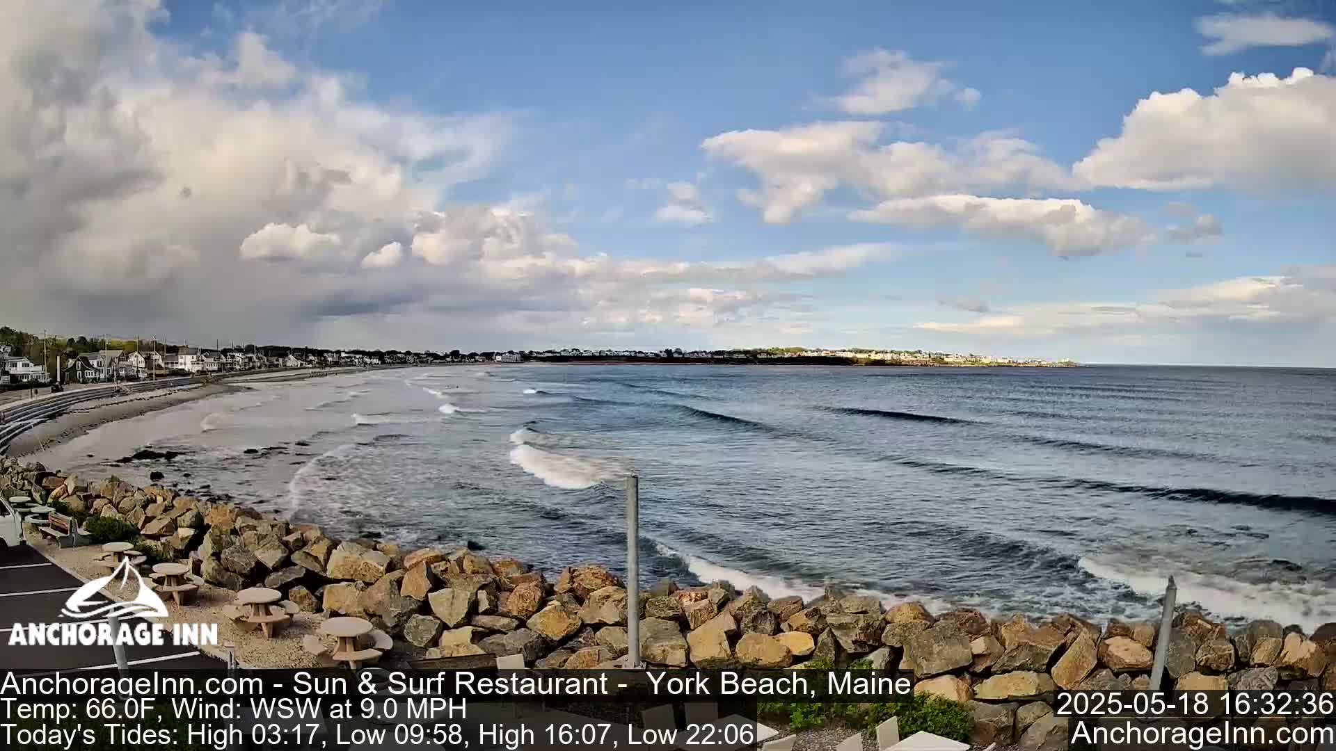 A partly cloudy day shows a sandy beach with small waves lapping a rocky shore, with a town visible in the distance.