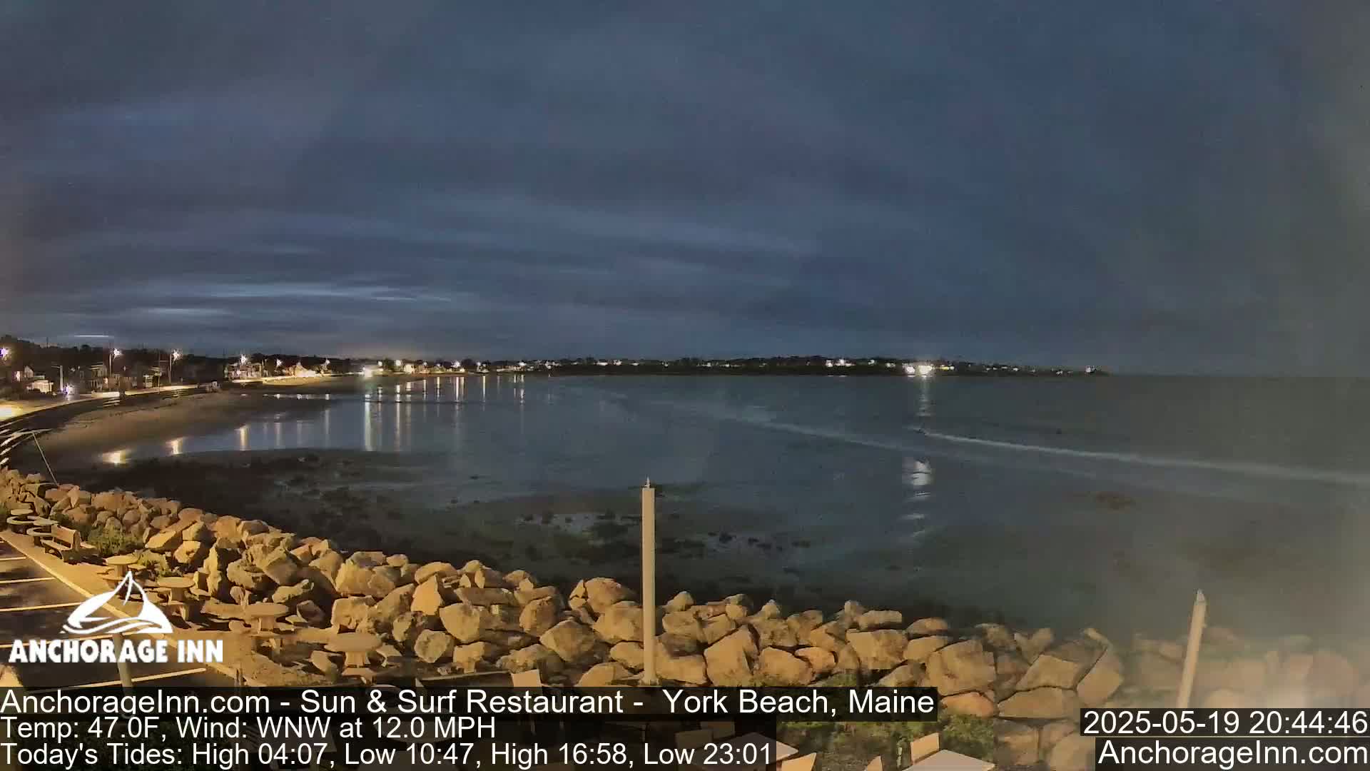 A nighttime coastal scene shows a calm body of water reflecting lights from a town across the bay, under a mostly clear, dark sky.