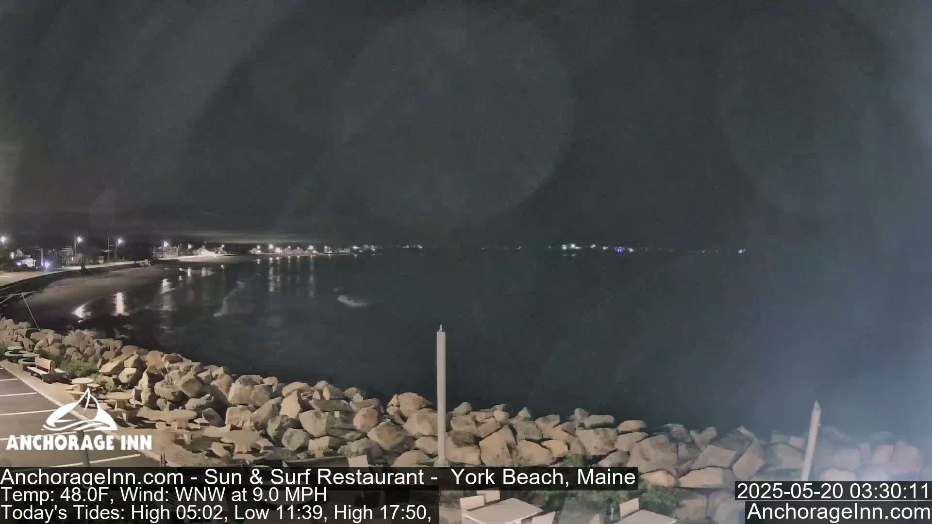 A nighttime view of a rocky shoreline and calm dark water, with faint lights visible across the water under a dark sky.