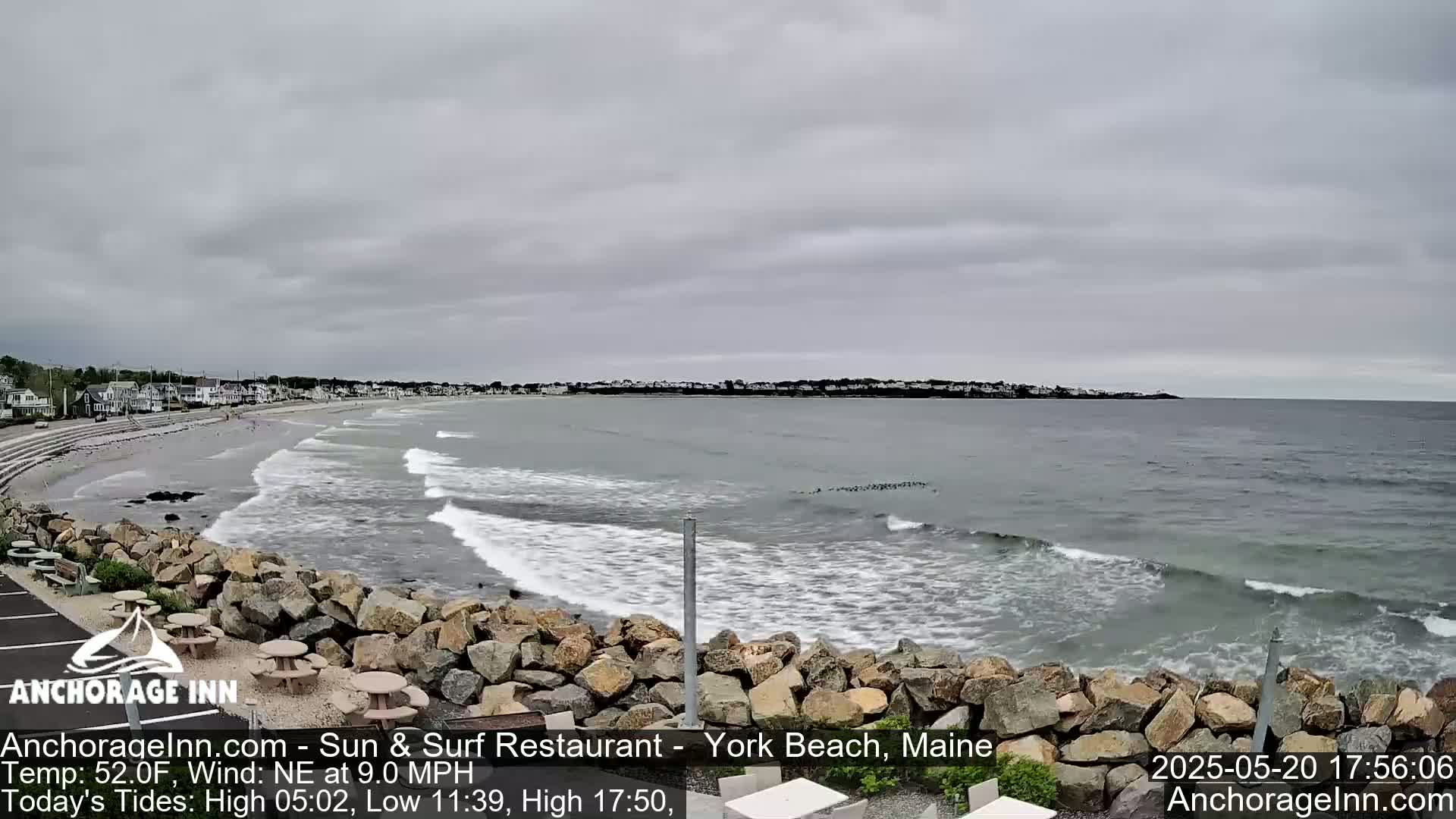 A view of a beach with gentle waves under an overcast sky, showing a rocky shoreline and distant houses.