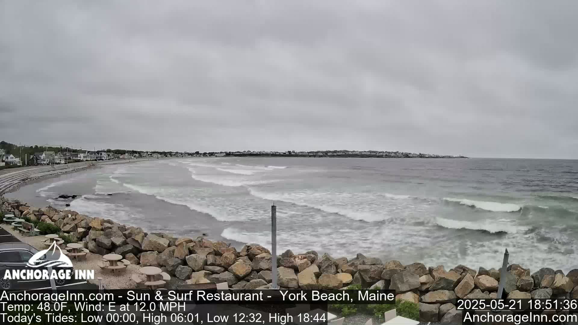 A beach with small waves under an overcast sky is viewed from a rocky shoreline with picnic tables.