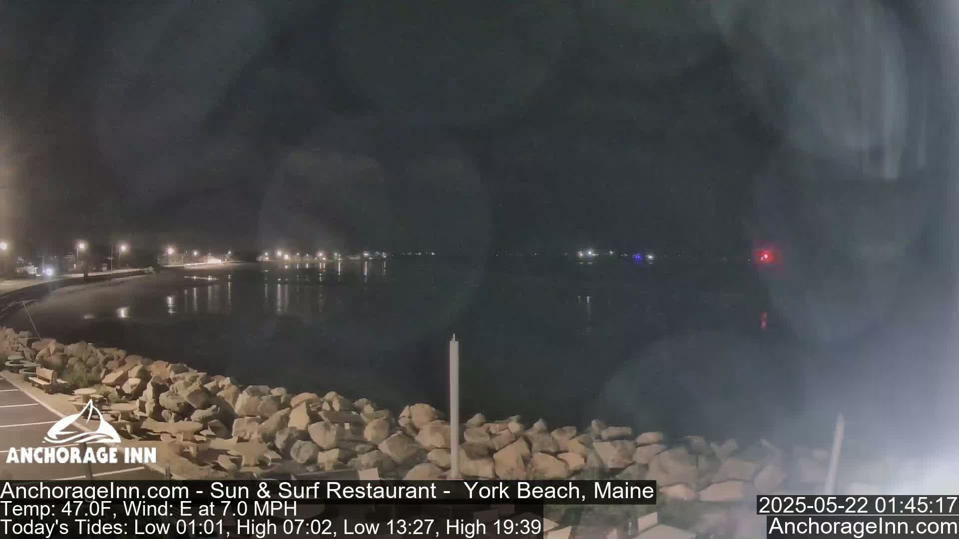 A nighttime view of a calm body of water, bordered by a rocky shoreline and dimly lit buildings in the distance, under a dark sky.