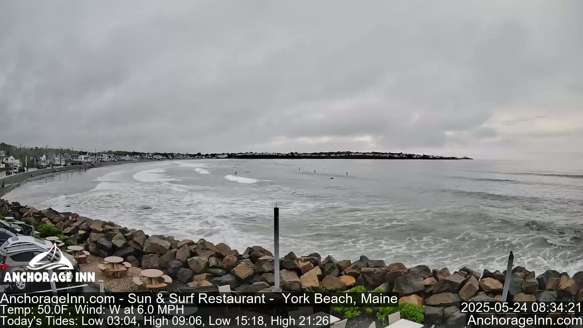 A rocky shoreline overlooks a grey, overcast ocean with several people surfing in the water.
