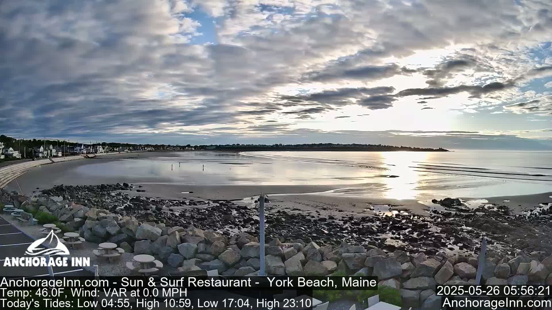 A mostly cloudy sunrise illuminates a beach with calm waves, rocky shoreline, and houses in the distance.