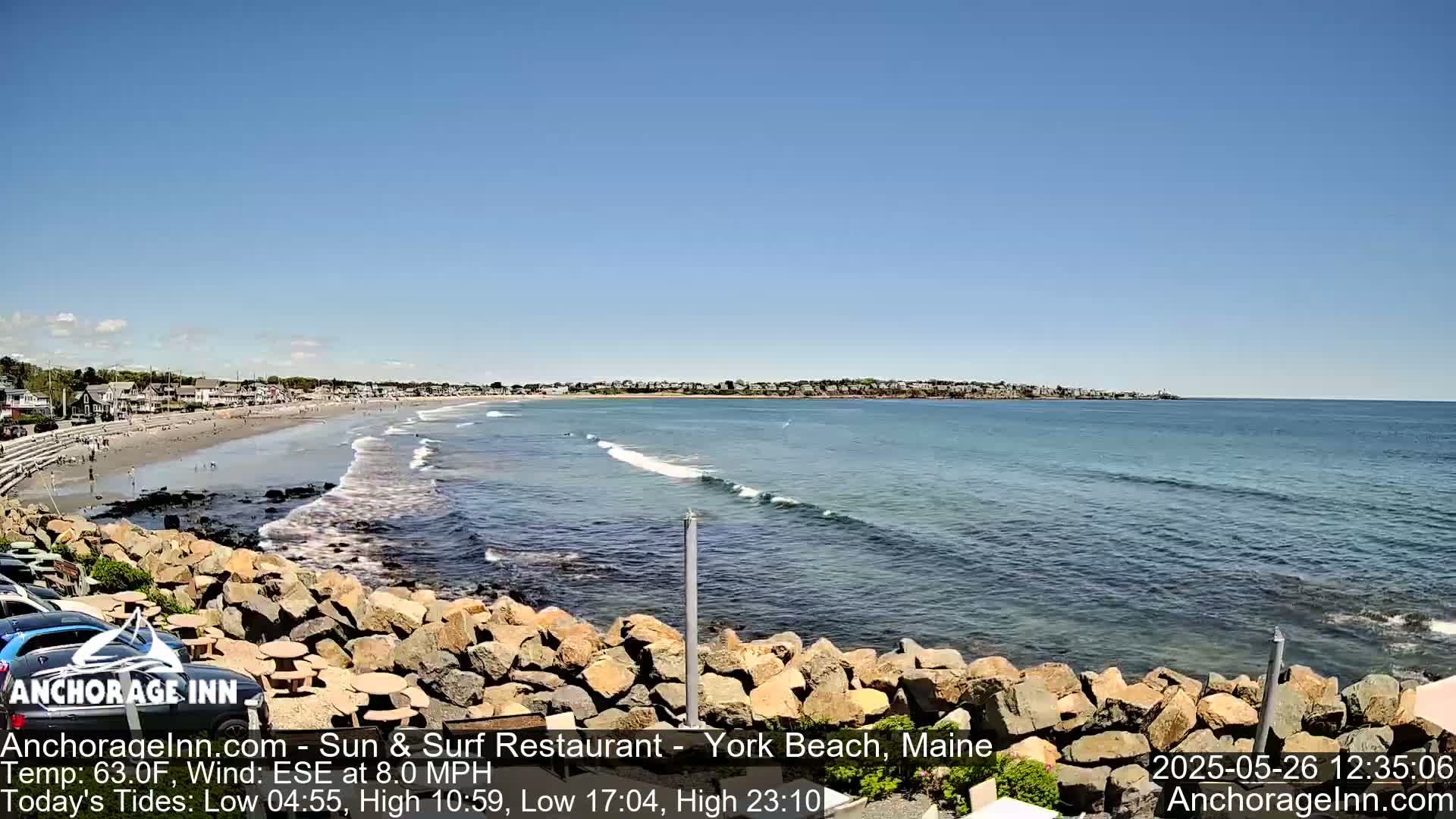 A sandy beach with gentle waves and a few people stretches along a coastline, under a clear blue sky.