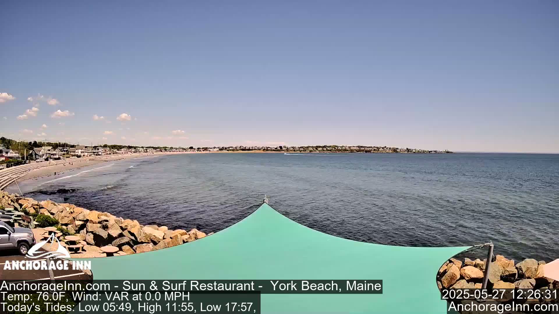A sandy beach with calm ocean water and a row of houses stretches across the horizon under a mostly sunny sky.