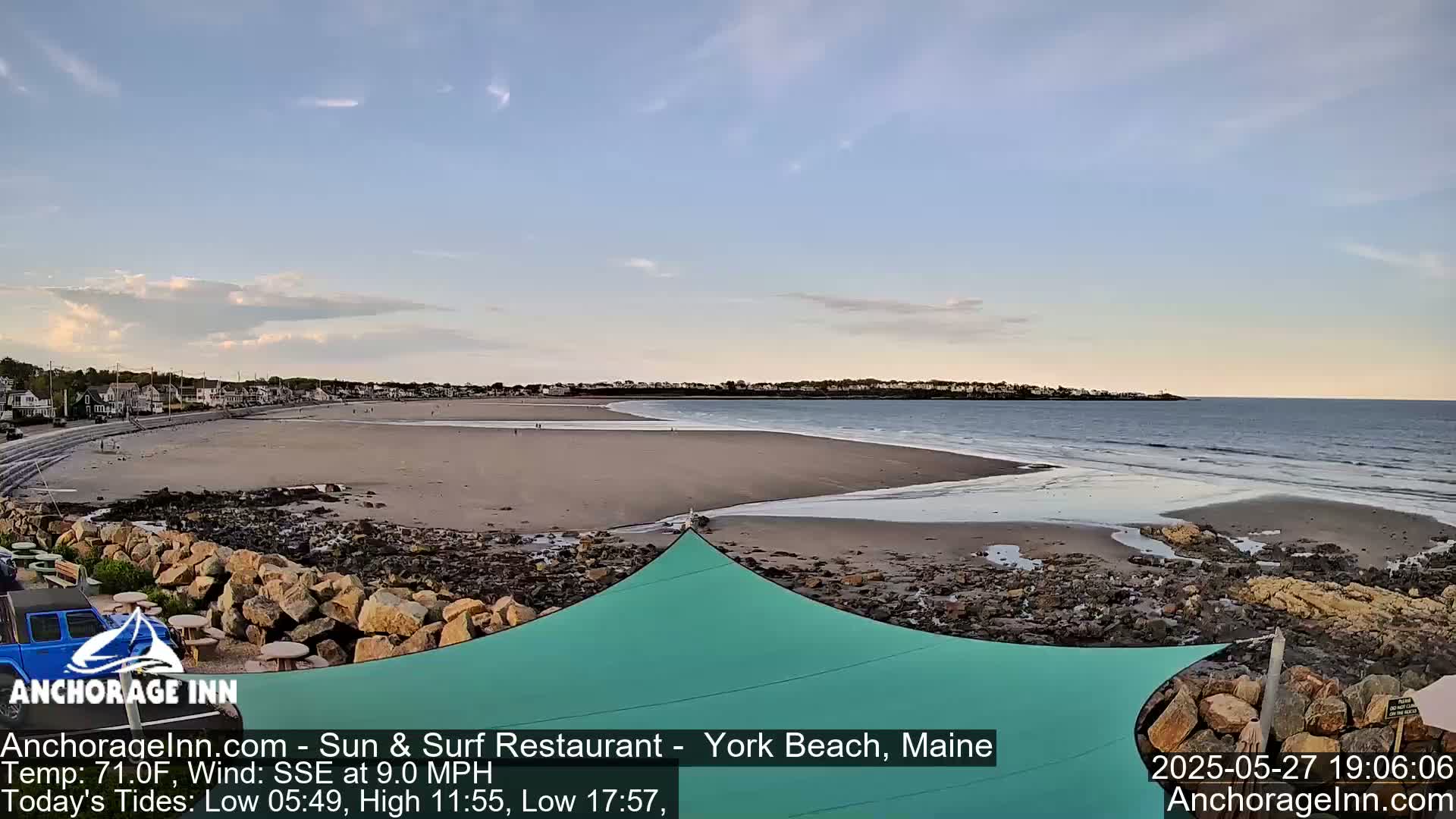 A sandy beach with calm waves under a partly cloudy sky is viewed from a rocky shoreline, partially shaded by a teal fabric shade structure.