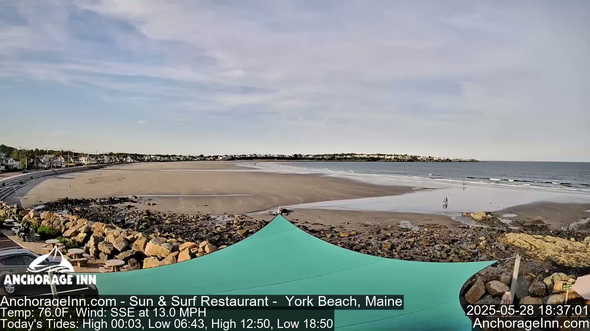 A sandy beach with a teal sunshade in the foreground stretches out to a calm ocean under a mostly clear sky.