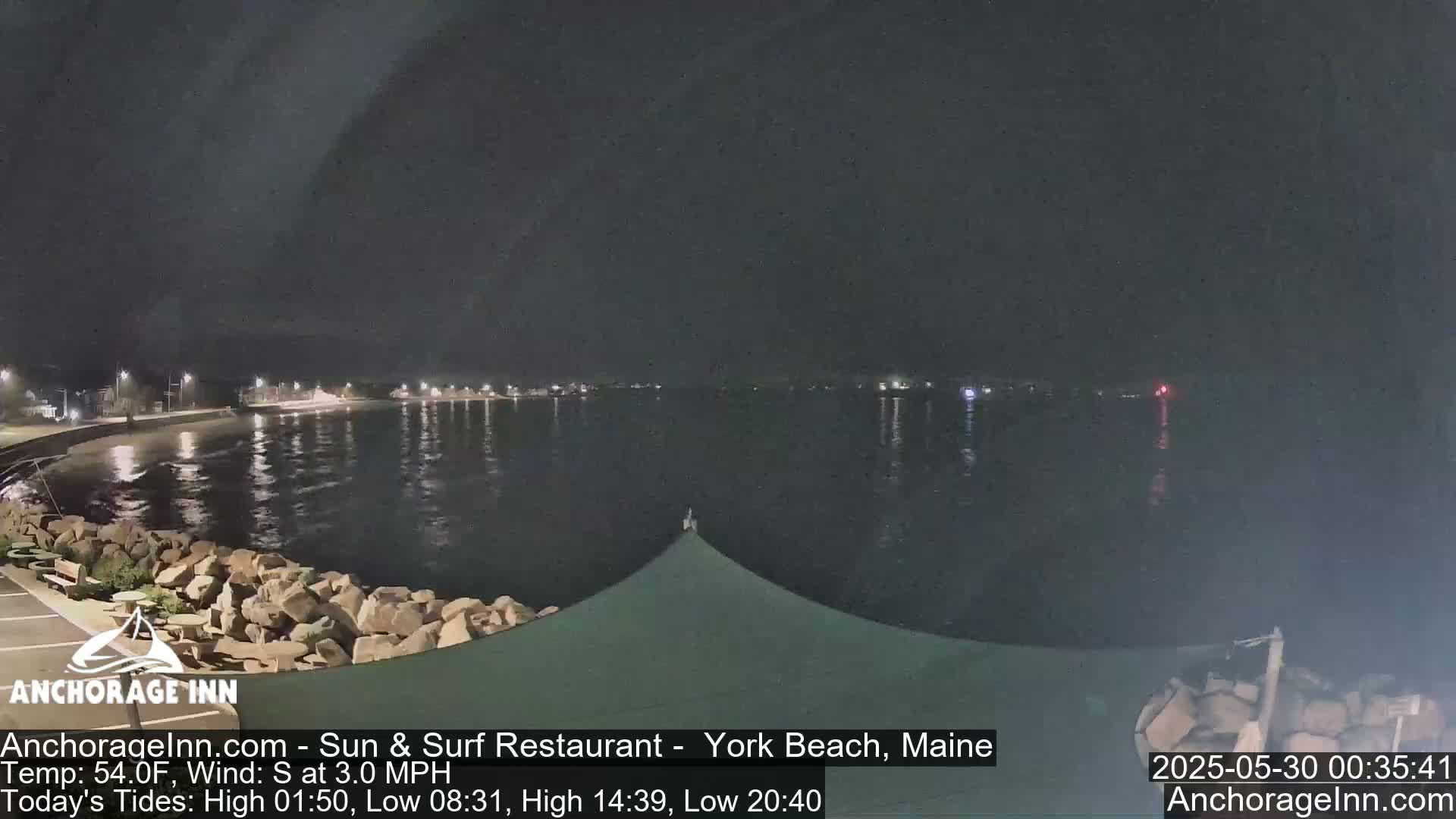 A nighttime view of a calm, dark body of water with a shoreline featuring rocks and a partially visible structure under a dark sky.