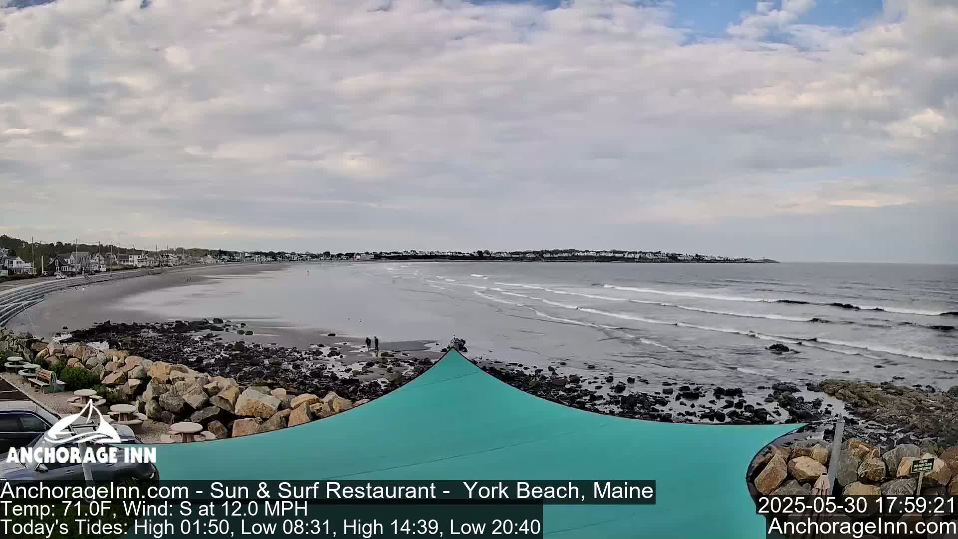 A partly cloudy day at a beach shows a wide sandy shore with small waves rolling in, a teal shade structure, and a rocky shoreline.