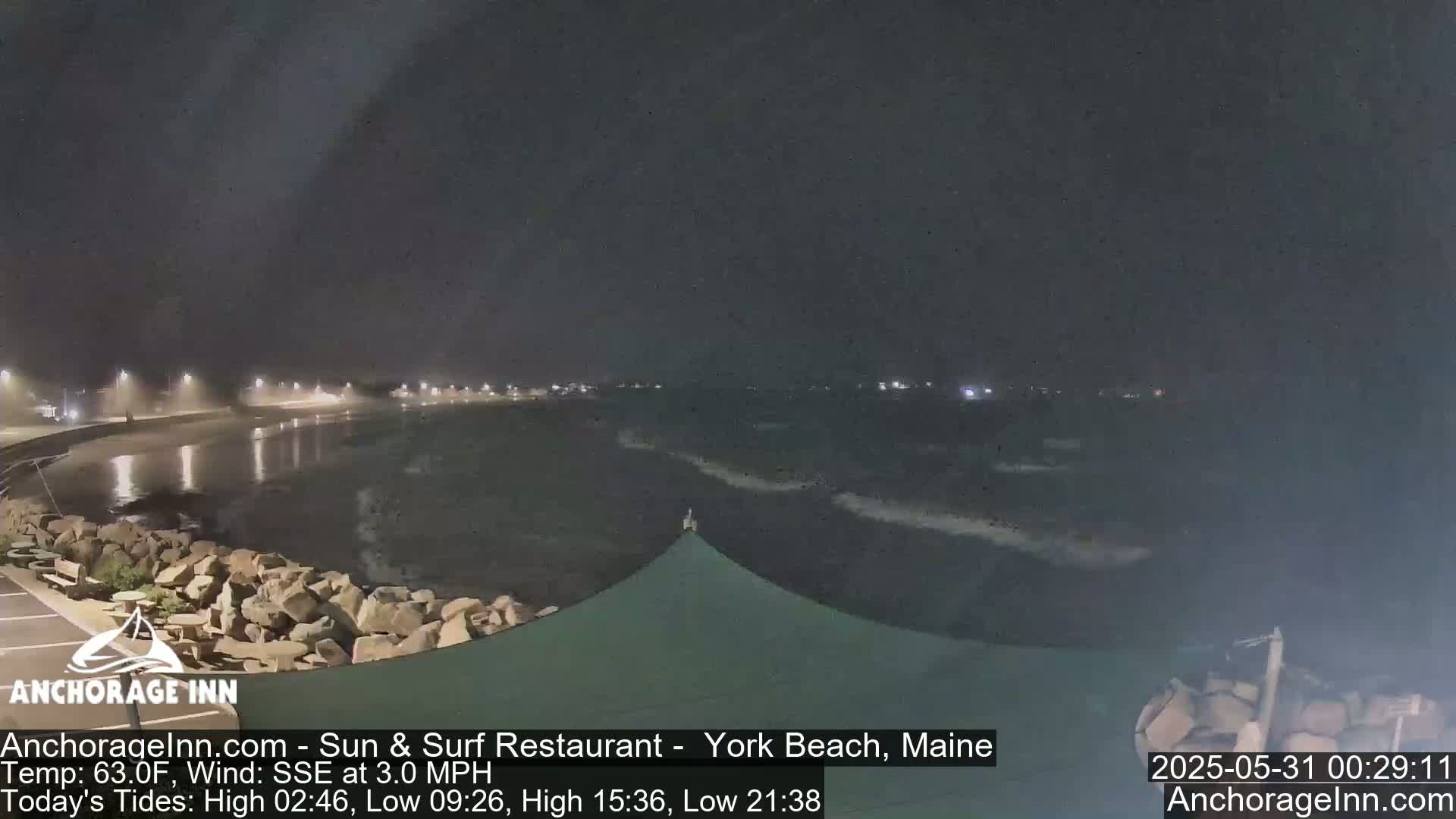 A nighttime view of a dark, wavy ocean under a cloudy sky, with a rocky shoreline and dimly lit buildings in the distance.