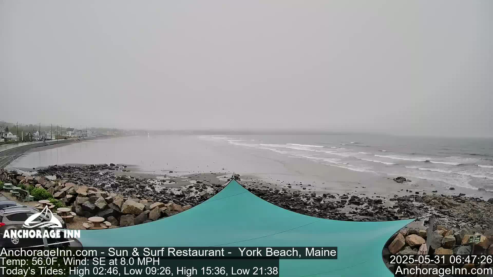 A hazy, overcast day at a rocky beach shows a calm sea with small waves, a teal shade sail, and a few buildings visible in the distance.