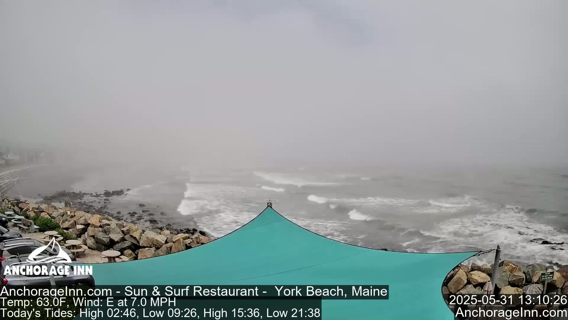 A foggy coastal scene shows a rocky shoreline with a teal shade structure, and a gray ocean with small waves in the distance.