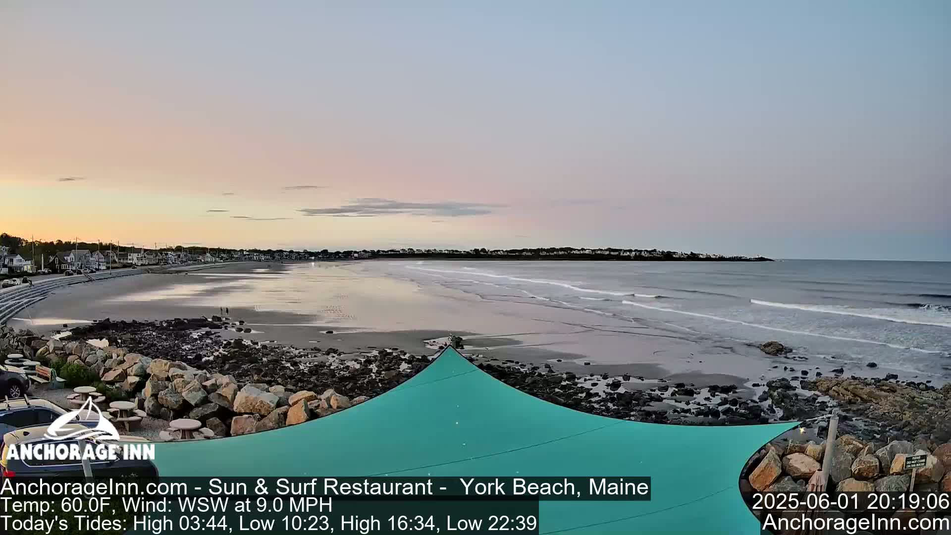 A tranquil beach scene at sunset shows gentle waves lapping the shore, with a calm ocean and residential buildings in the background under a pastel sky.