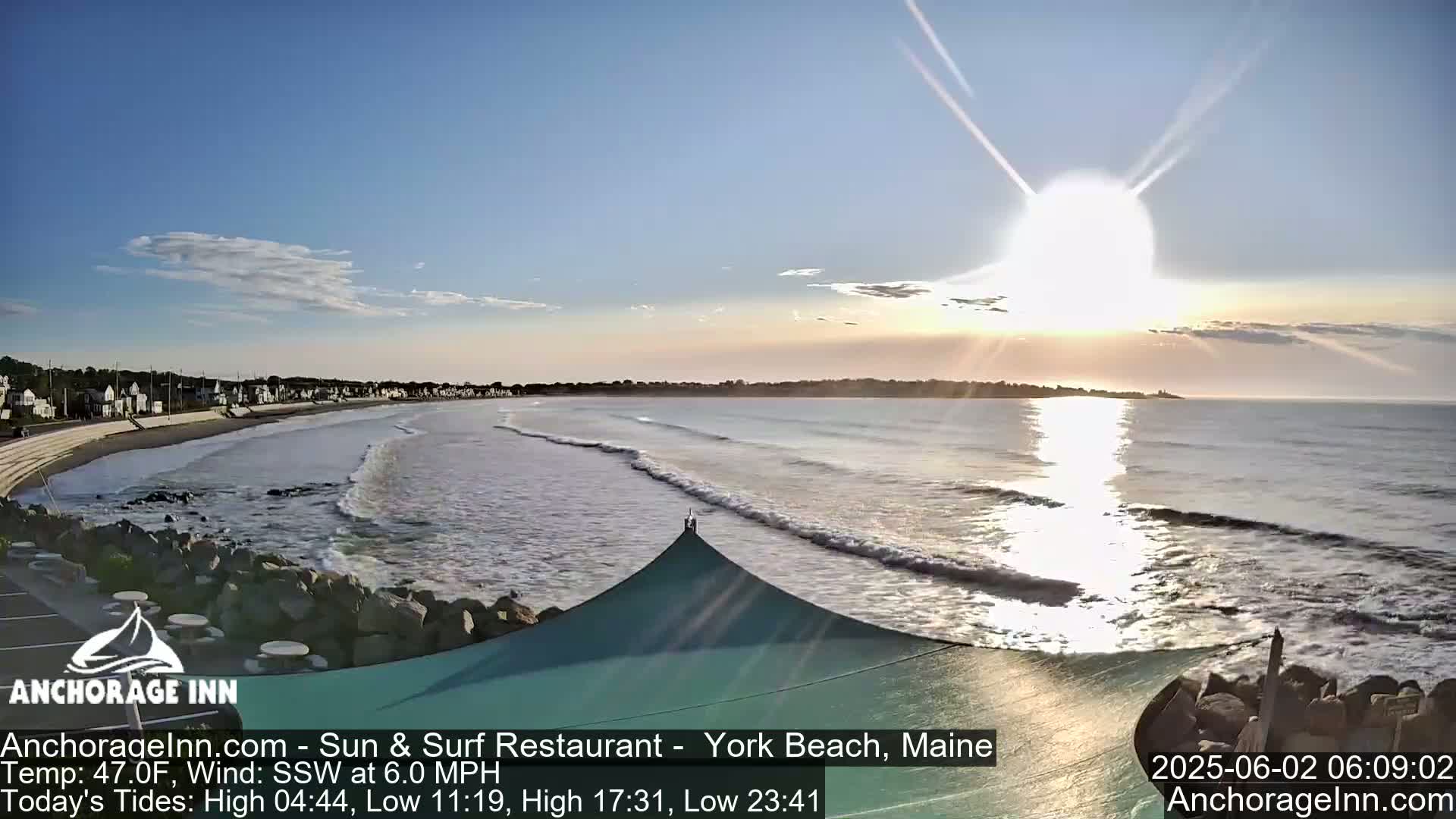 A sunny morning reveals a beach with gentle waves lapping the shore,  a town visible in the distance, and a partially covered patio in the foreground.