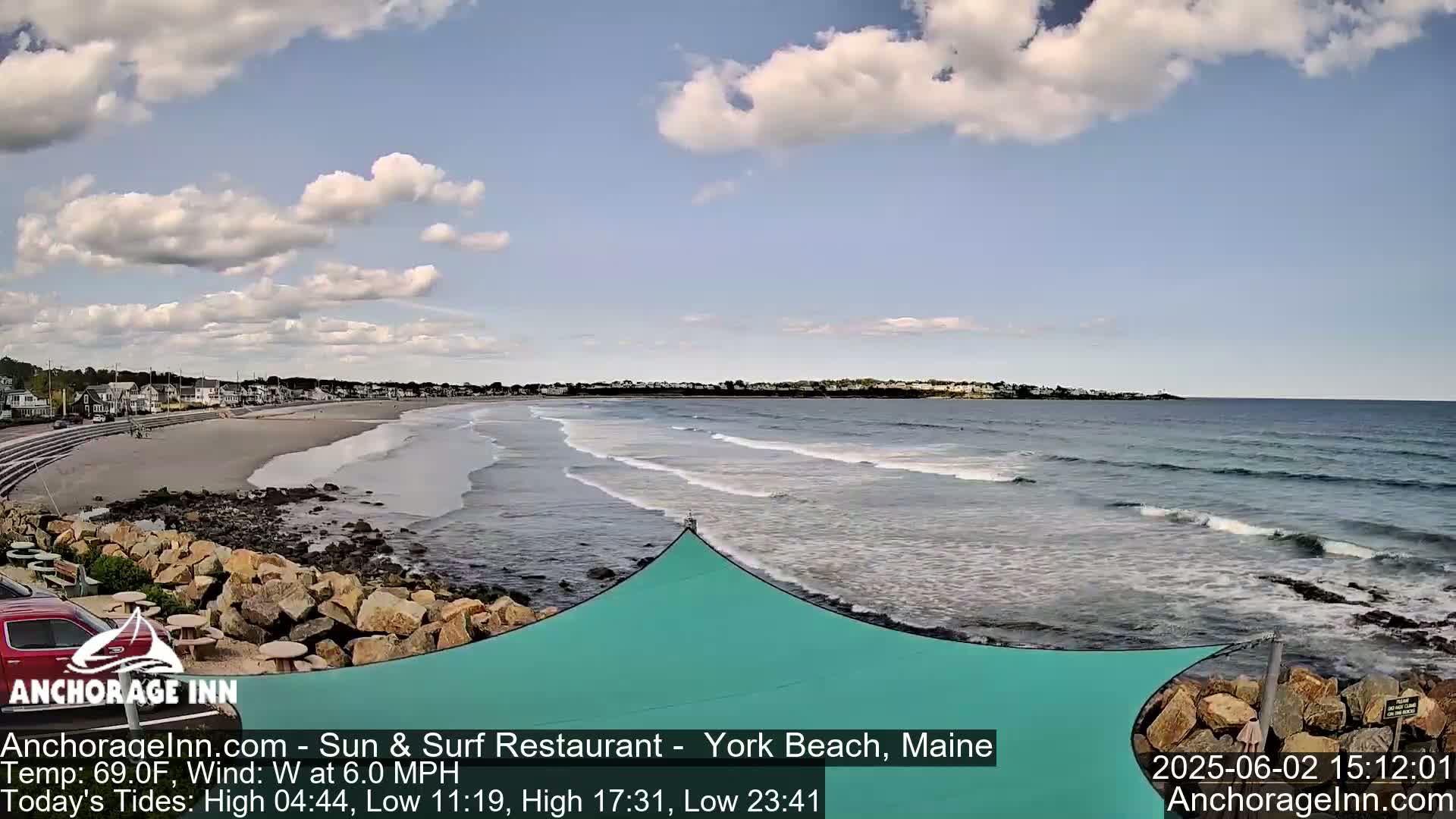 A sandy beach with small waves under a partly cloudy sky, viewed from a rocky shore with a teal shade structure.