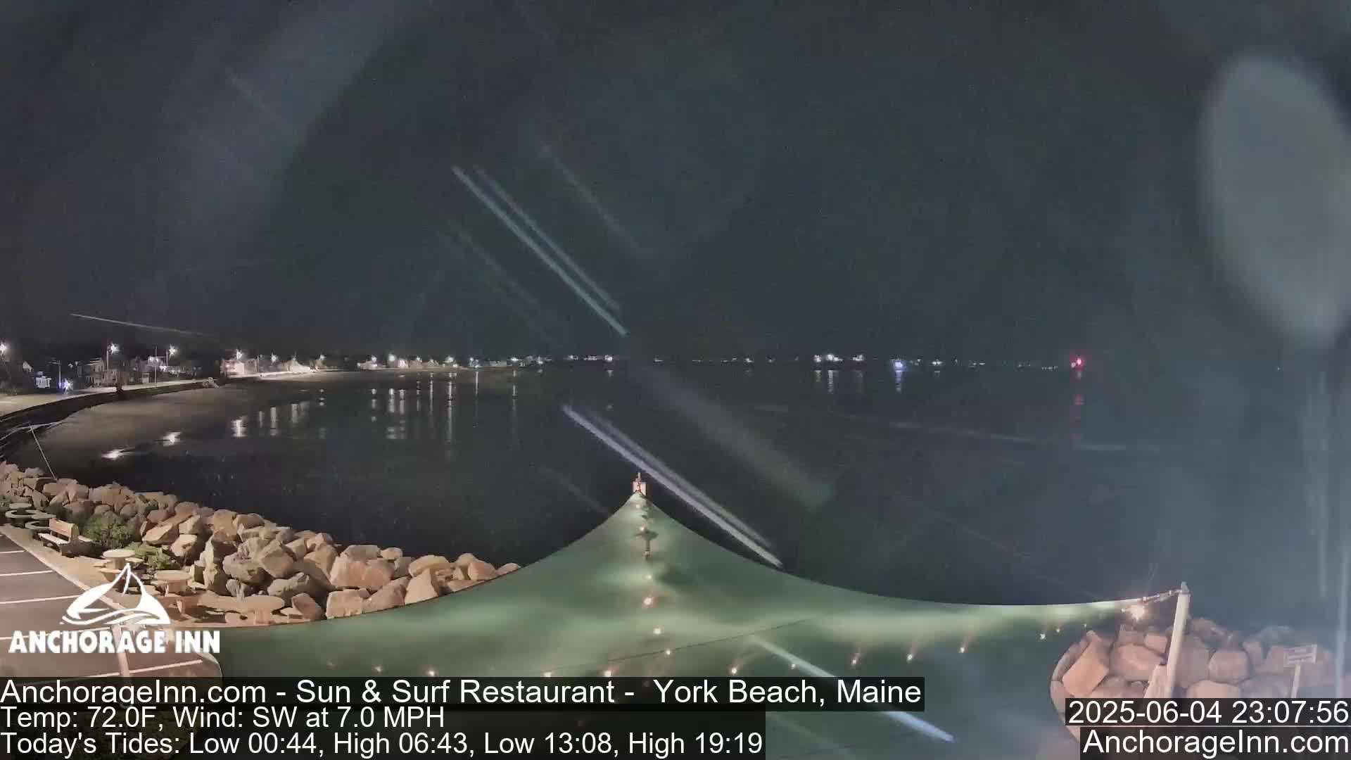 A nighttime view of a calm body of water next to a rocky shoreline, with a partially visible structure and lights from buildings in the distance.