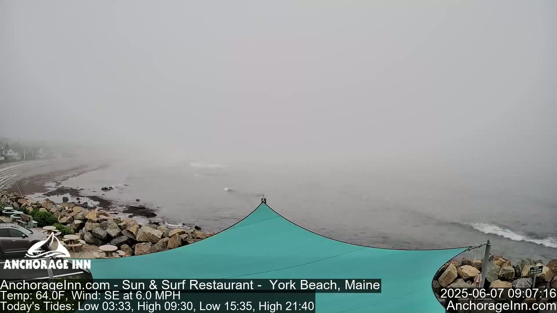 A foggy beach scene shows a rocky shoreline, a teal-colored shade structure, and a calm ocean under an overcast sky.