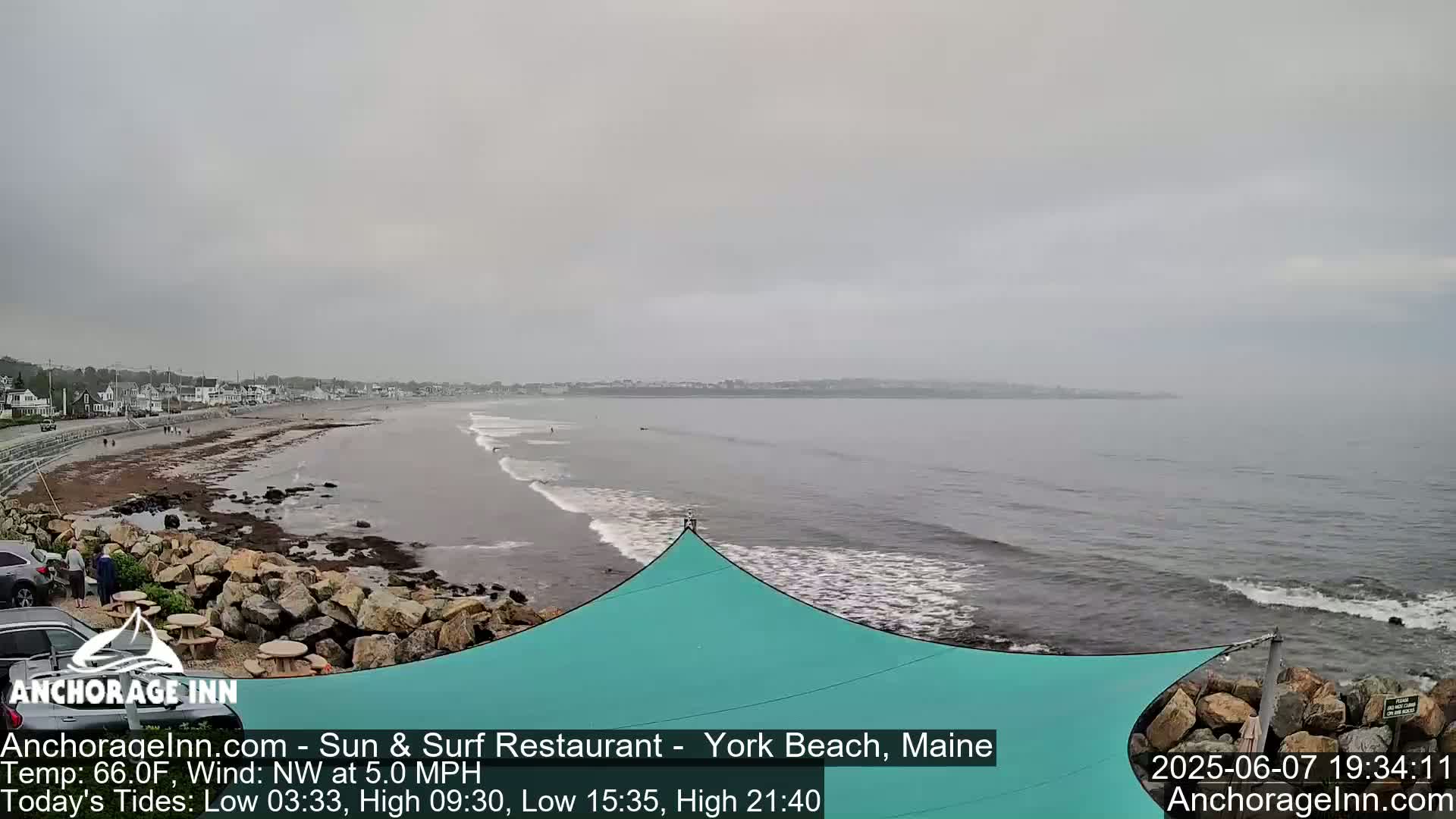 A view of a beach with overcast skies, gentle waves lapping the shore, and a teal shade structure in the foreground.