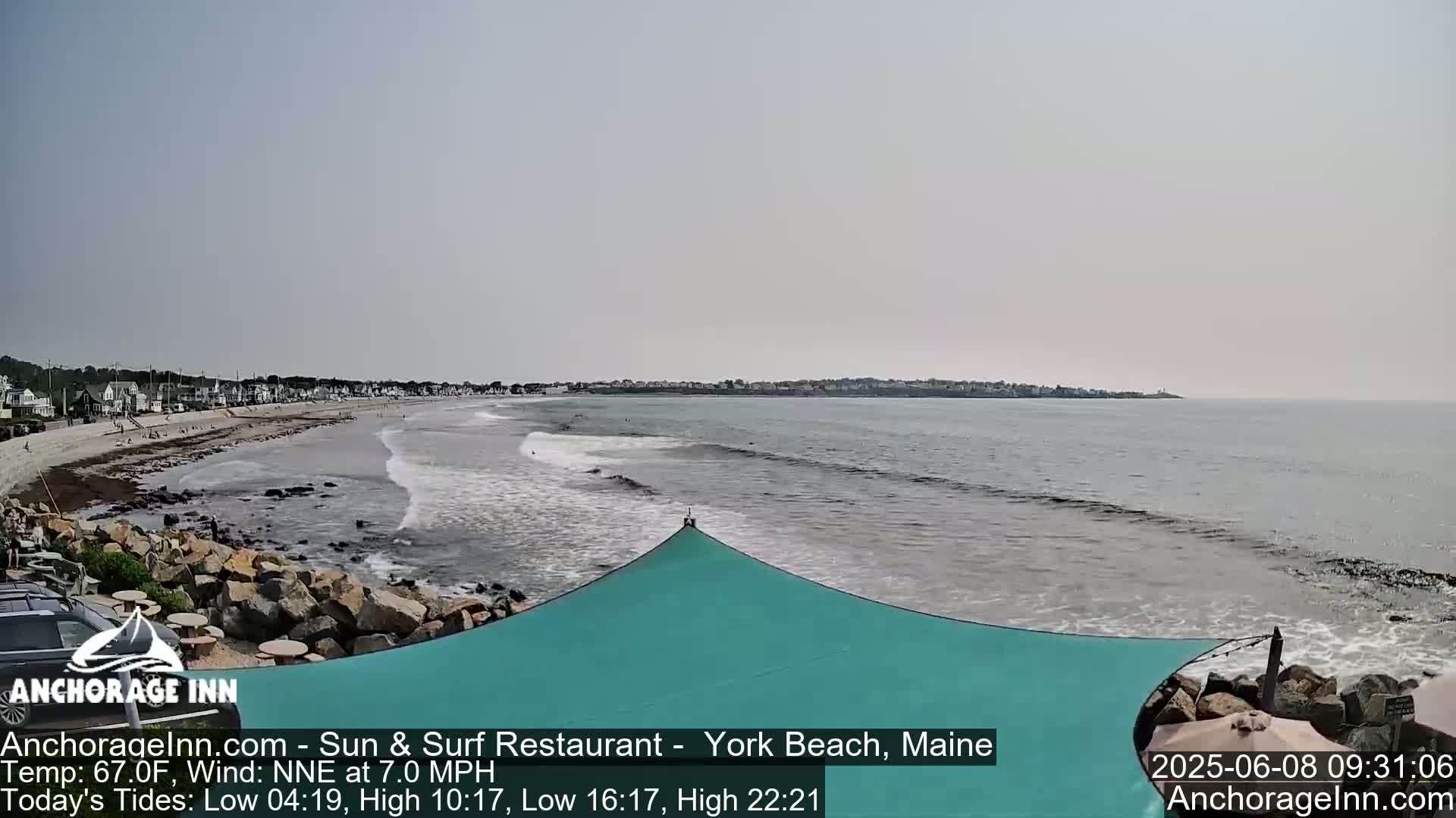 A sandy beach with calm waves and a teal sunshade overlooks a town under a clear sky.
