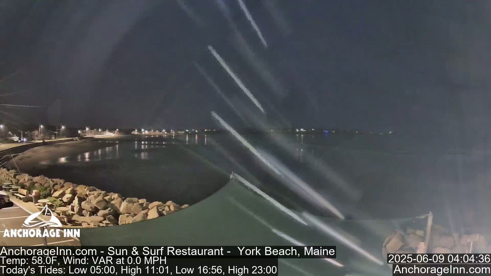 A nighttime view of a calm body of water next to a rocky shoreline, with faint streaks of light in the sky.
