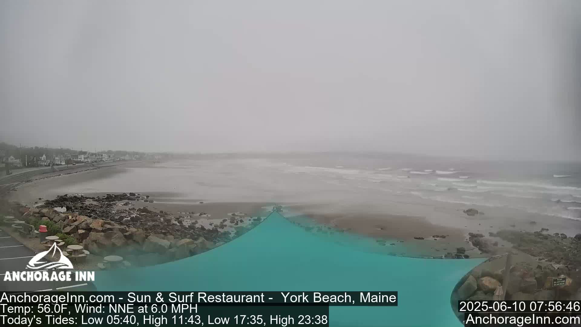 A foggy beach scene shows a calm ocean with small waves lapping a sandy shore near some buildings.
