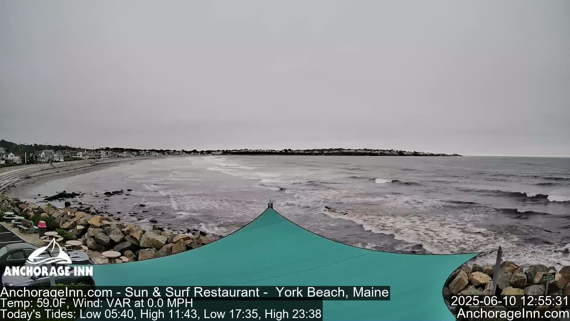 A mostly cloudy day shows a beach with a rocky shoreline, calm ocean waves, and a teal-colored shade structure.
