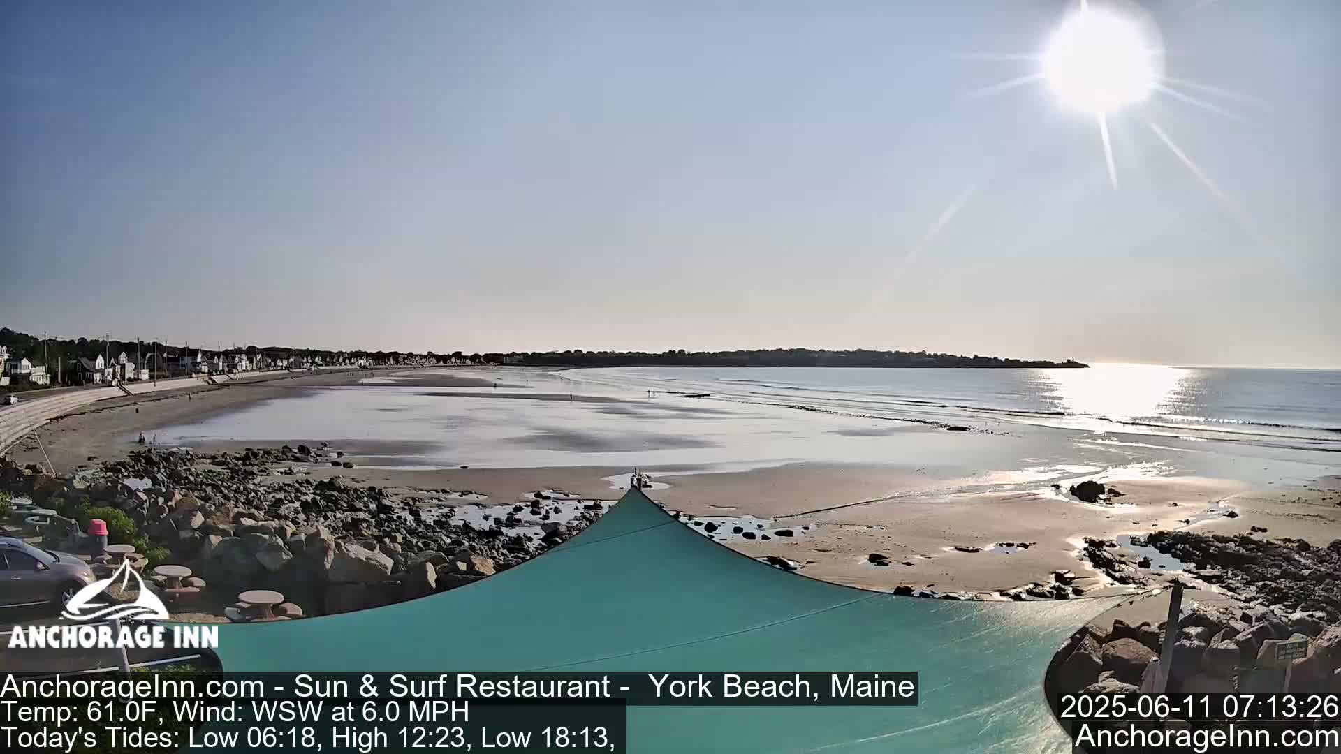 A sunny beach scene shows a wide expanse of sand, a calm ocean with small waves, and houses along the shoreline.