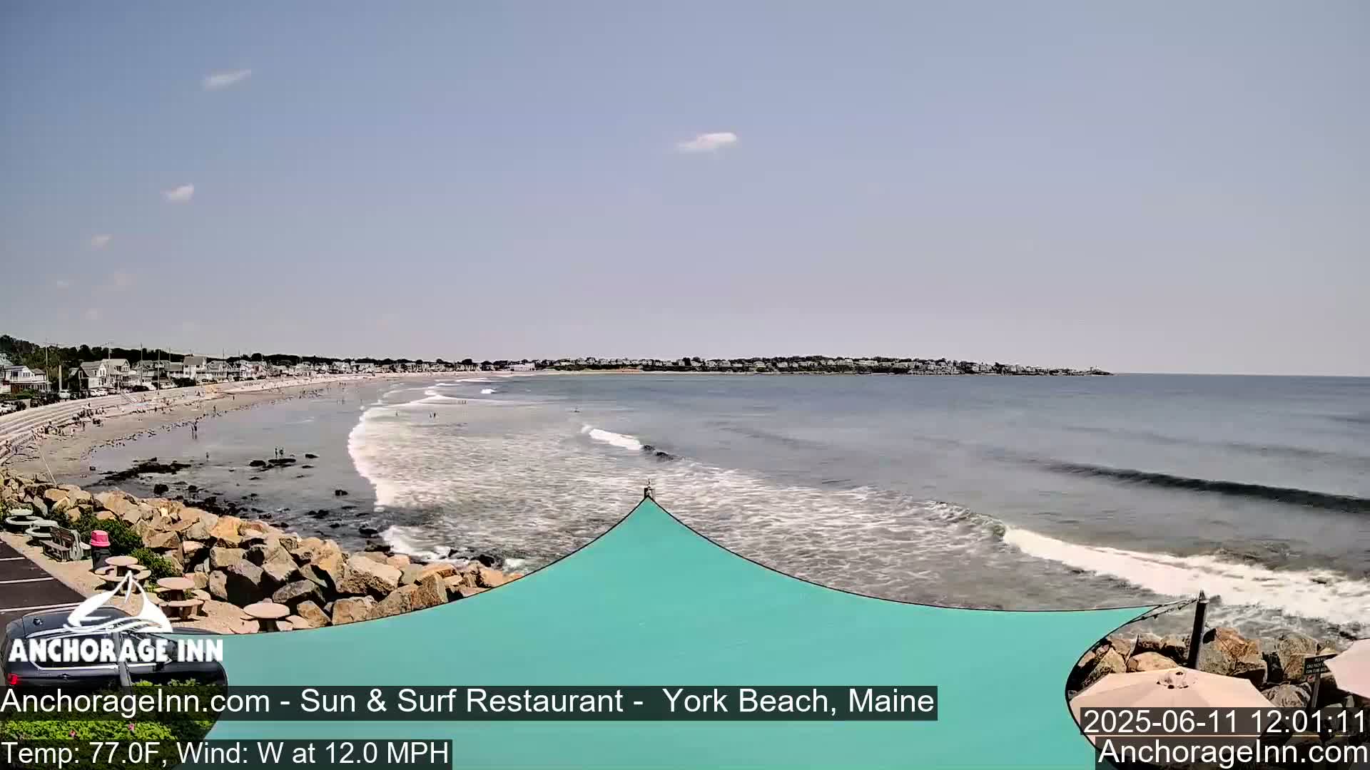 A sandy beach with a calm ocean and small waves under a mostly clear sky, showing a seaside town in the background.