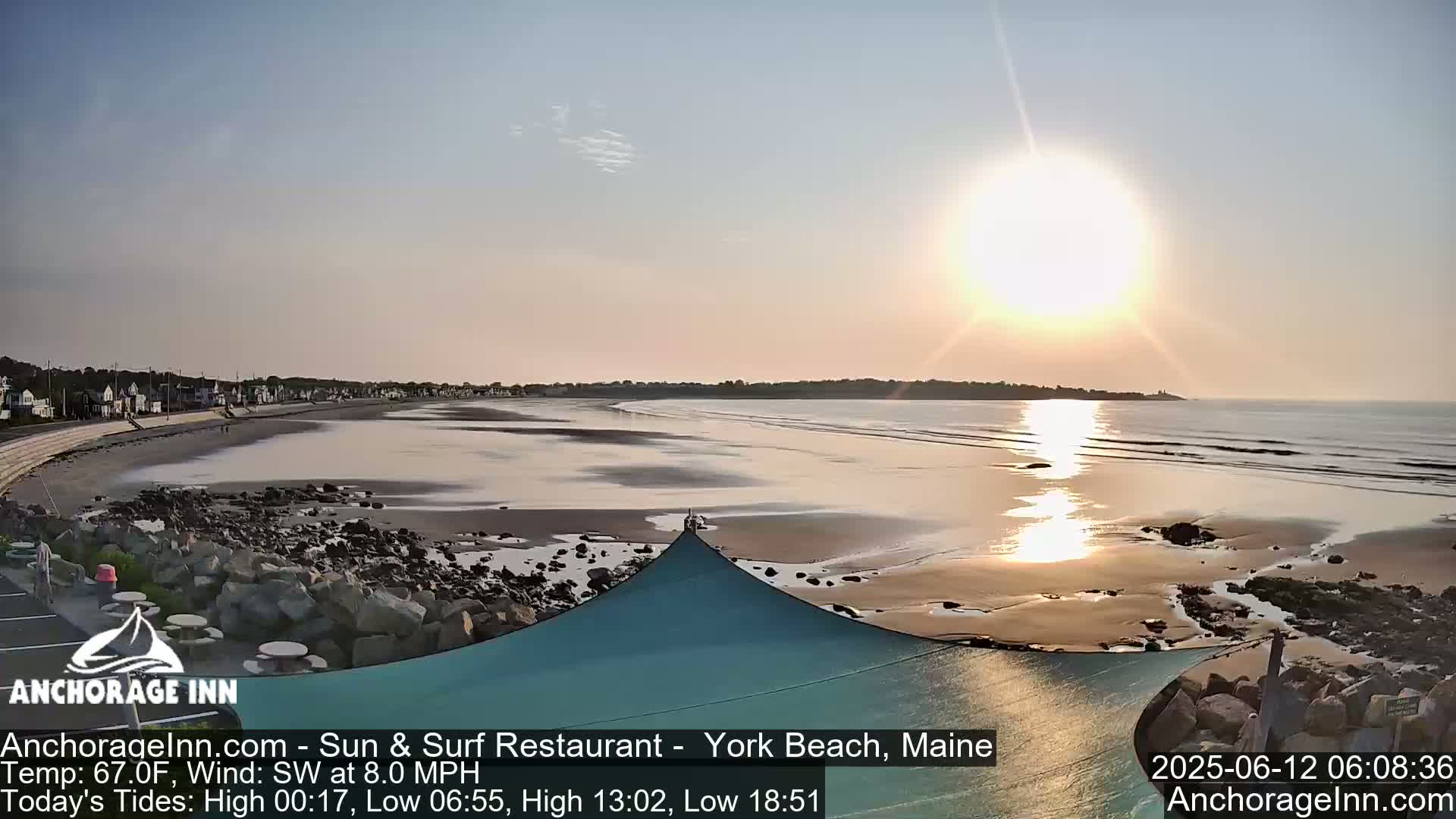 A sandy beach with calm waves stretches out to a bright sunrise over the ocean, with a small town visible in the distance.