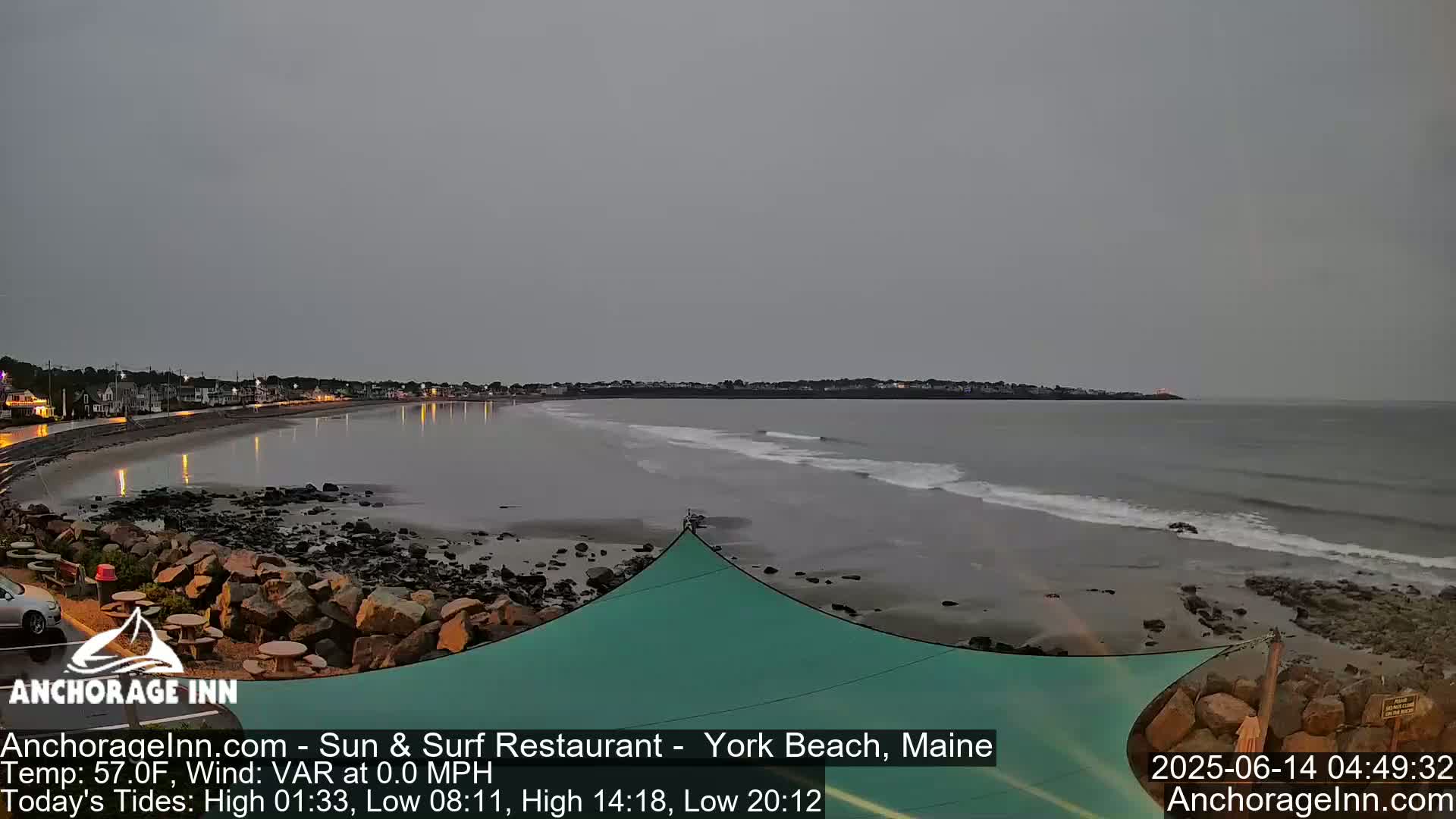 A beach scene under an overcast sky shows a calm ocean with small waves lapping the shore near a rocky coastline and a town in the distance.
