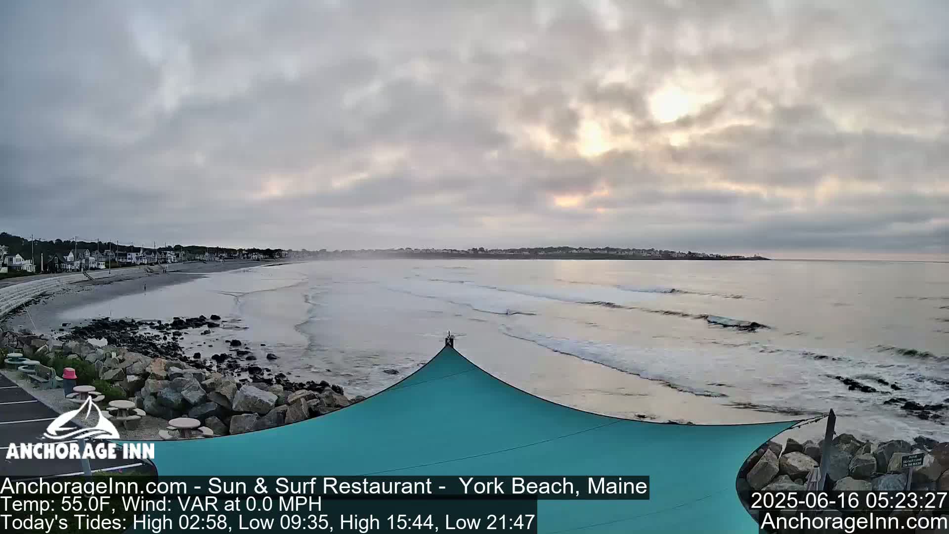 A mostly cloudy sunrise illuminates a beach with gentle waves lapping the shore, a teal shade structure partially visible in the foreground.