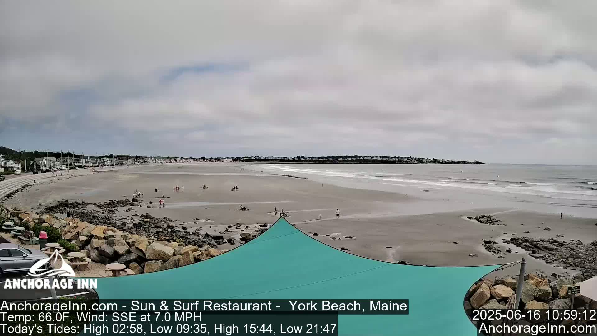 A wide sandy beach with a calm ocean under an overcast sky shows several people scattered along the shore near a teal shade structure.
