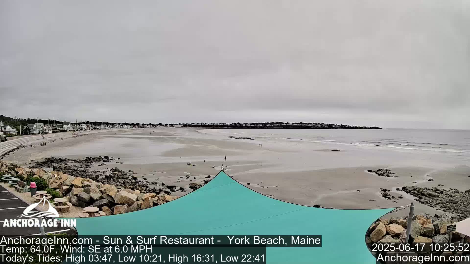 A wide sandy beach stretches to a calm ocean under an overcast sky, with a teal shade structure in the foreground and a town visible in the distance.