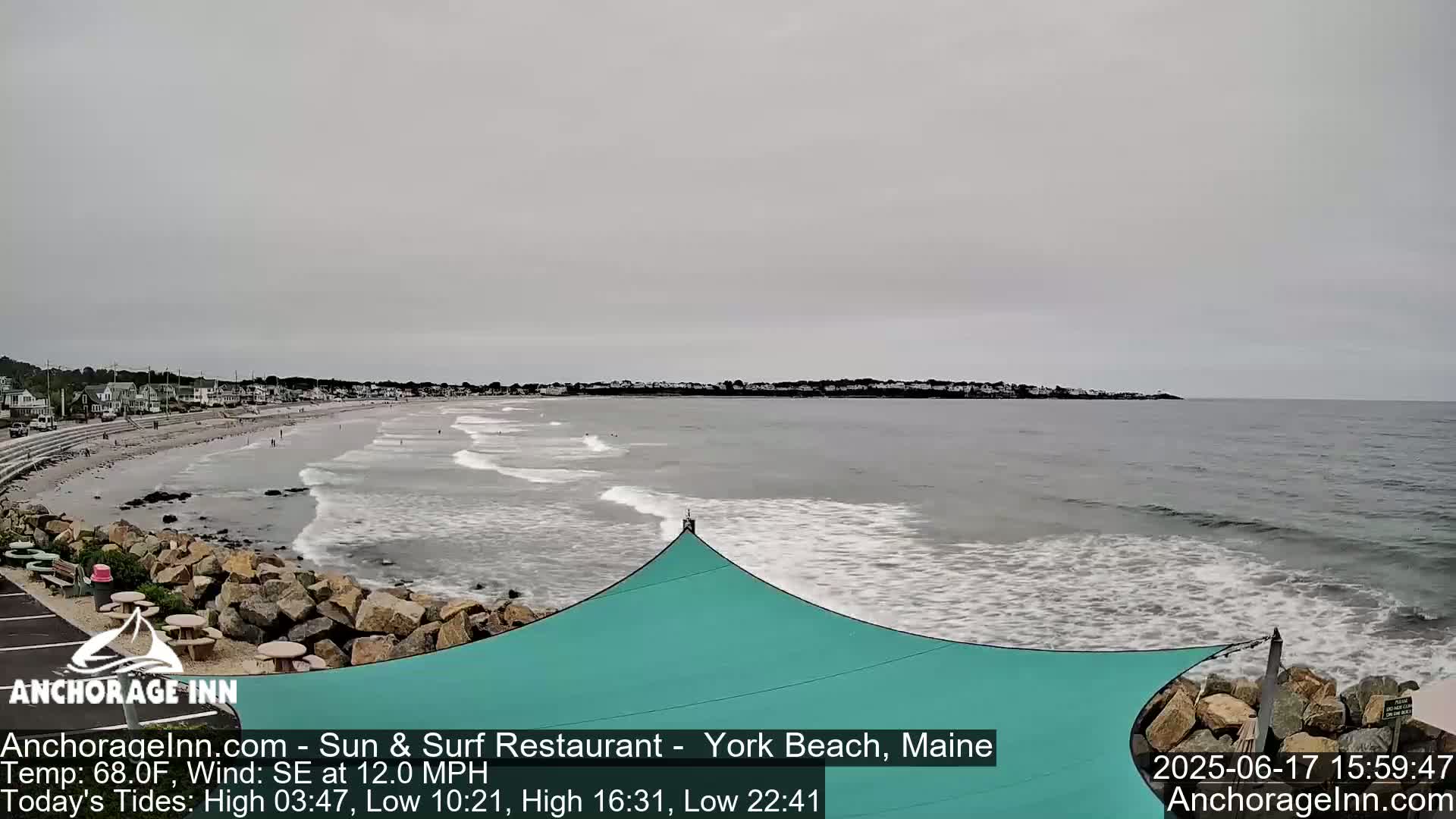 A sandy beach with small waves under an overcast sky, a teal shade structure is in the foreground next to a rocky outcropping.