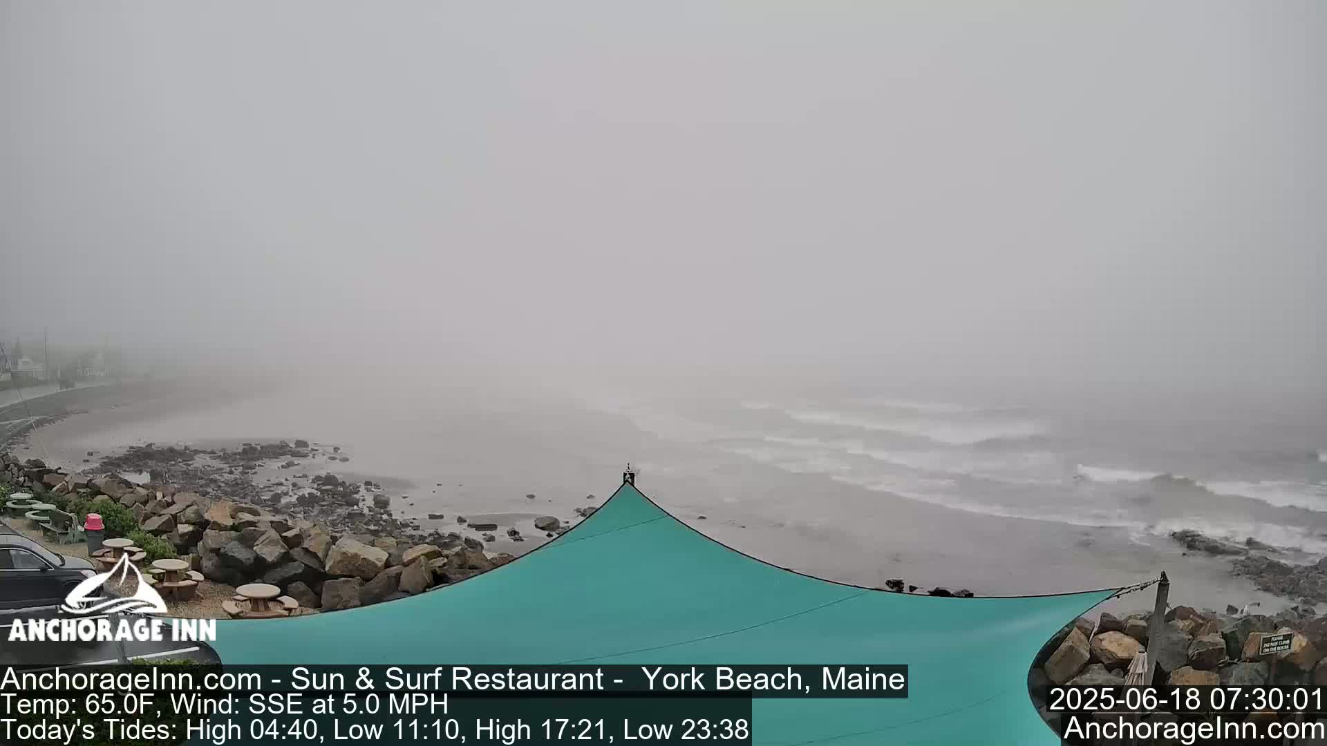 A foggy beach scene shows a rocky shoreline, small waves, and a teal-colored shade structure.
