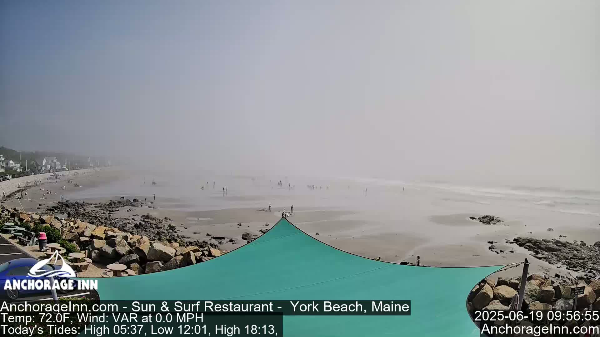 A hazy, calm beach scene shows a sandy shore with scattered people, a teal shade structure, and dark rocks near the water's edge.