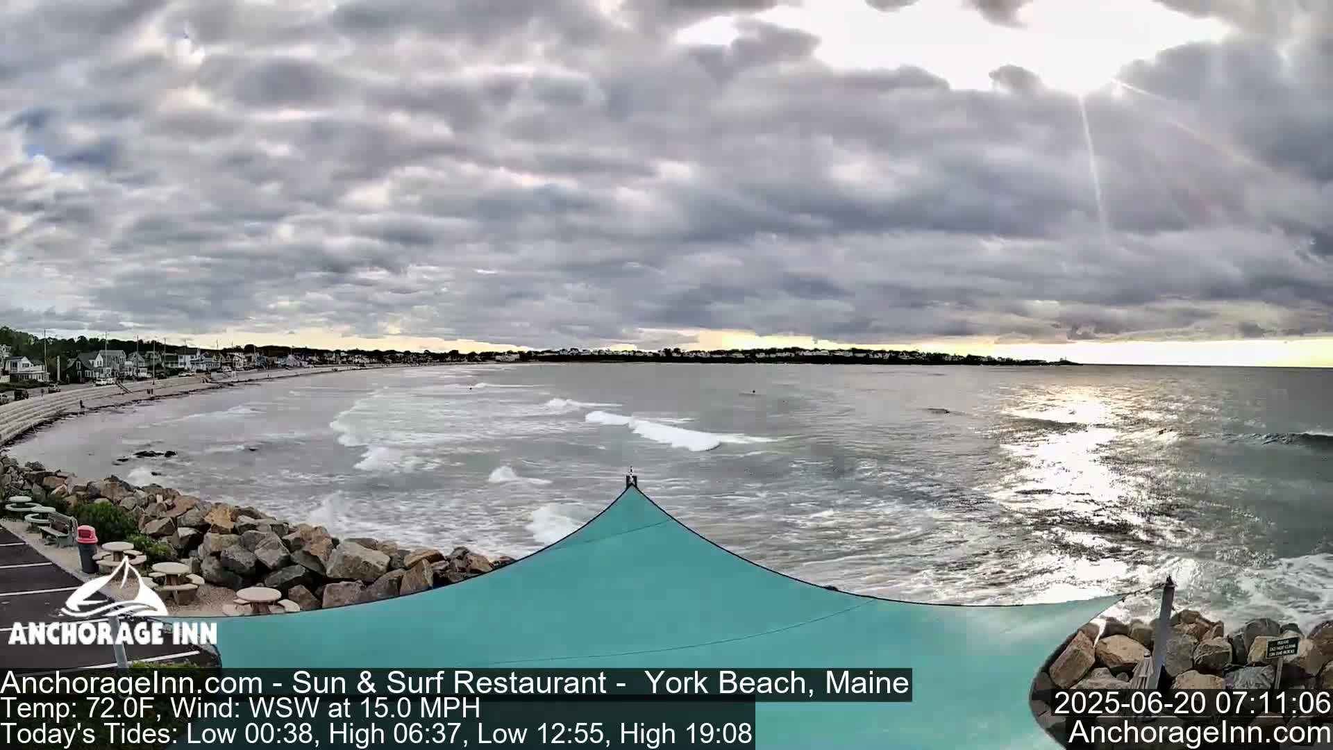 A mostly cloudy day at a beach with moderate waves and a teal-colored shade structure in the foreground.