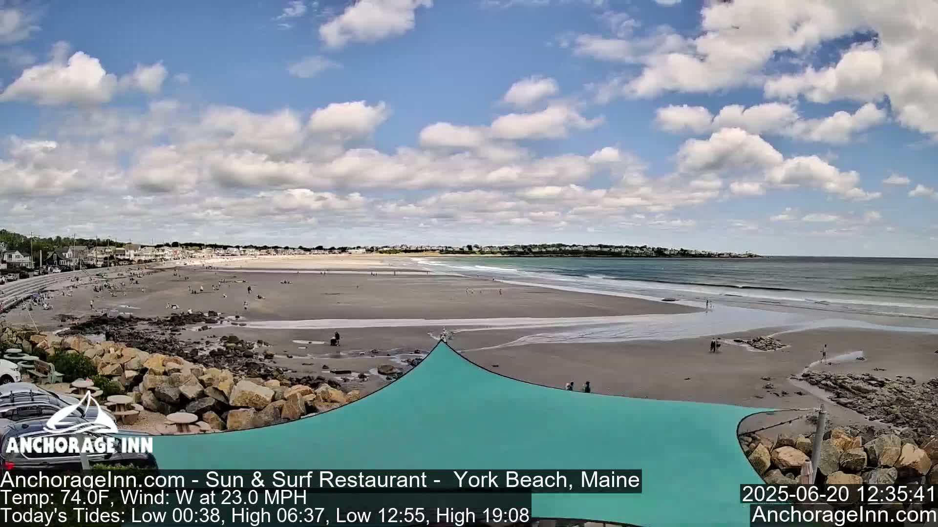 A sandy beach with a partly cloudy sky and a teal shade structure is seen from a high vantage point, showing a calm ocean with small waves and people enjoying the beach.